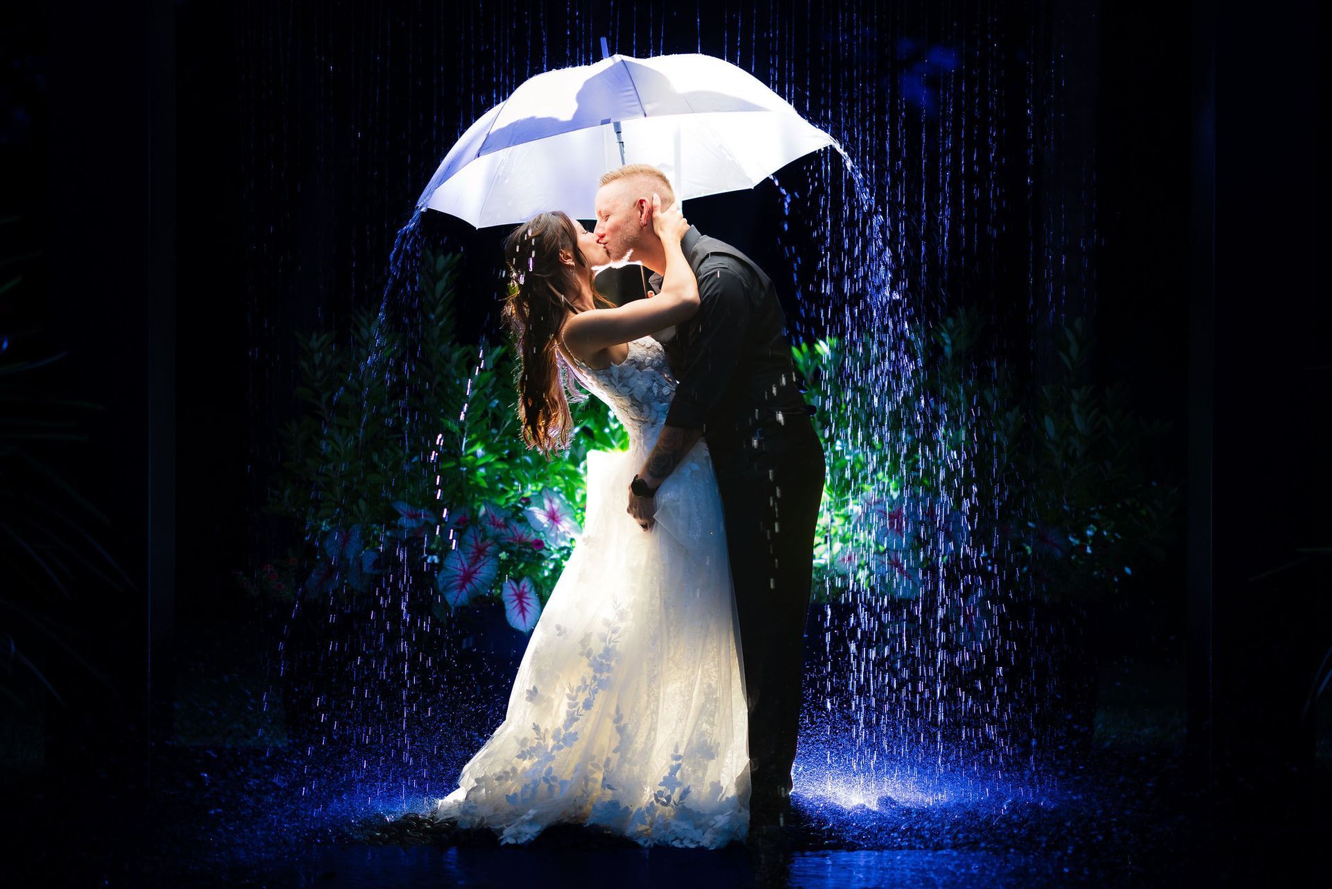 Bride and groom kissing under a white umbrella in the rain, lit by blue and green lights