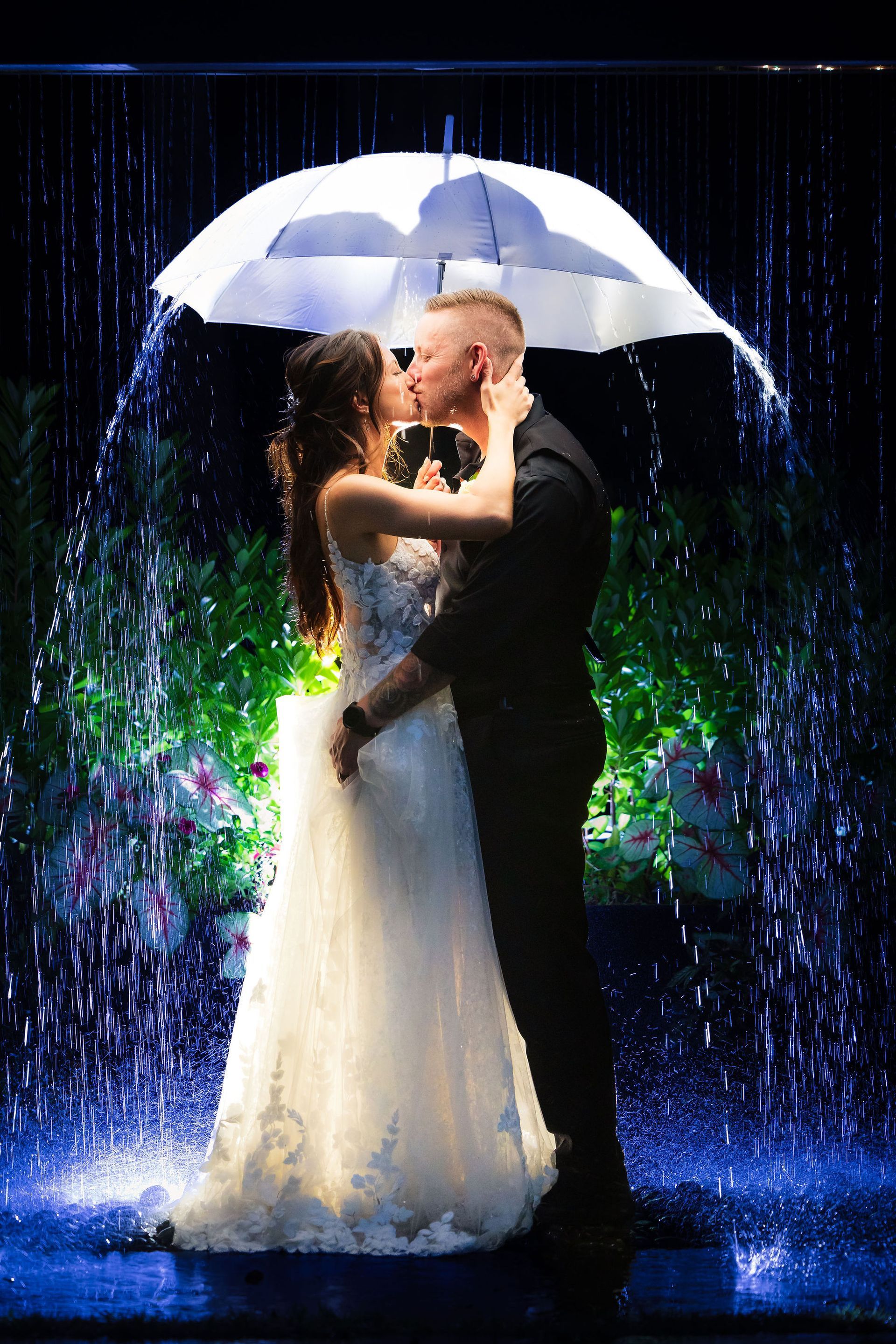 Bride and groom embrace under a white umbrella in a rain-lit nighttime scene