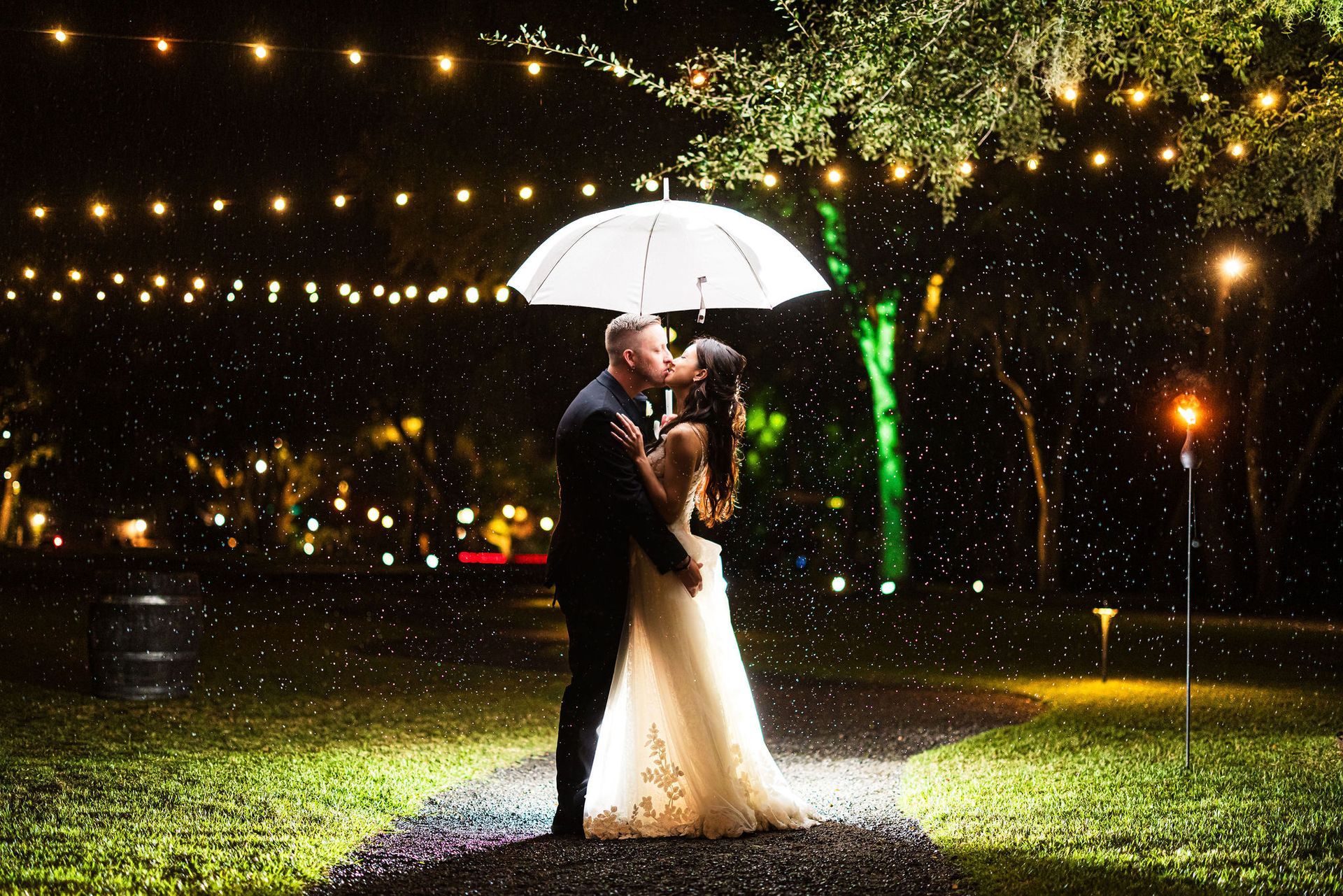 Couple embracing under a white umbrella in a rainy night garden, with warm lights glowing in the background
