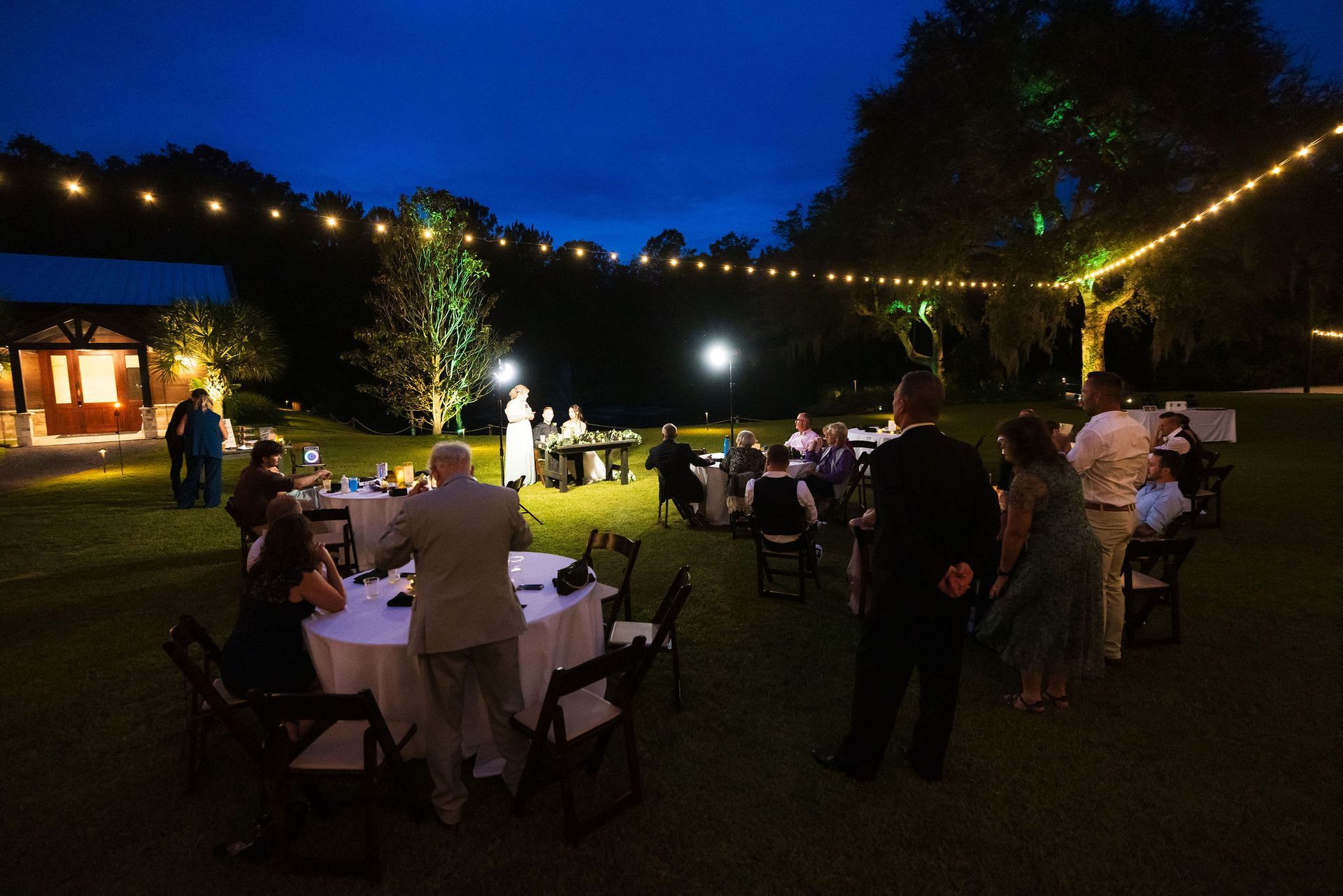 People seated at round tables in a garden at night, with string lights and a lit stage.
