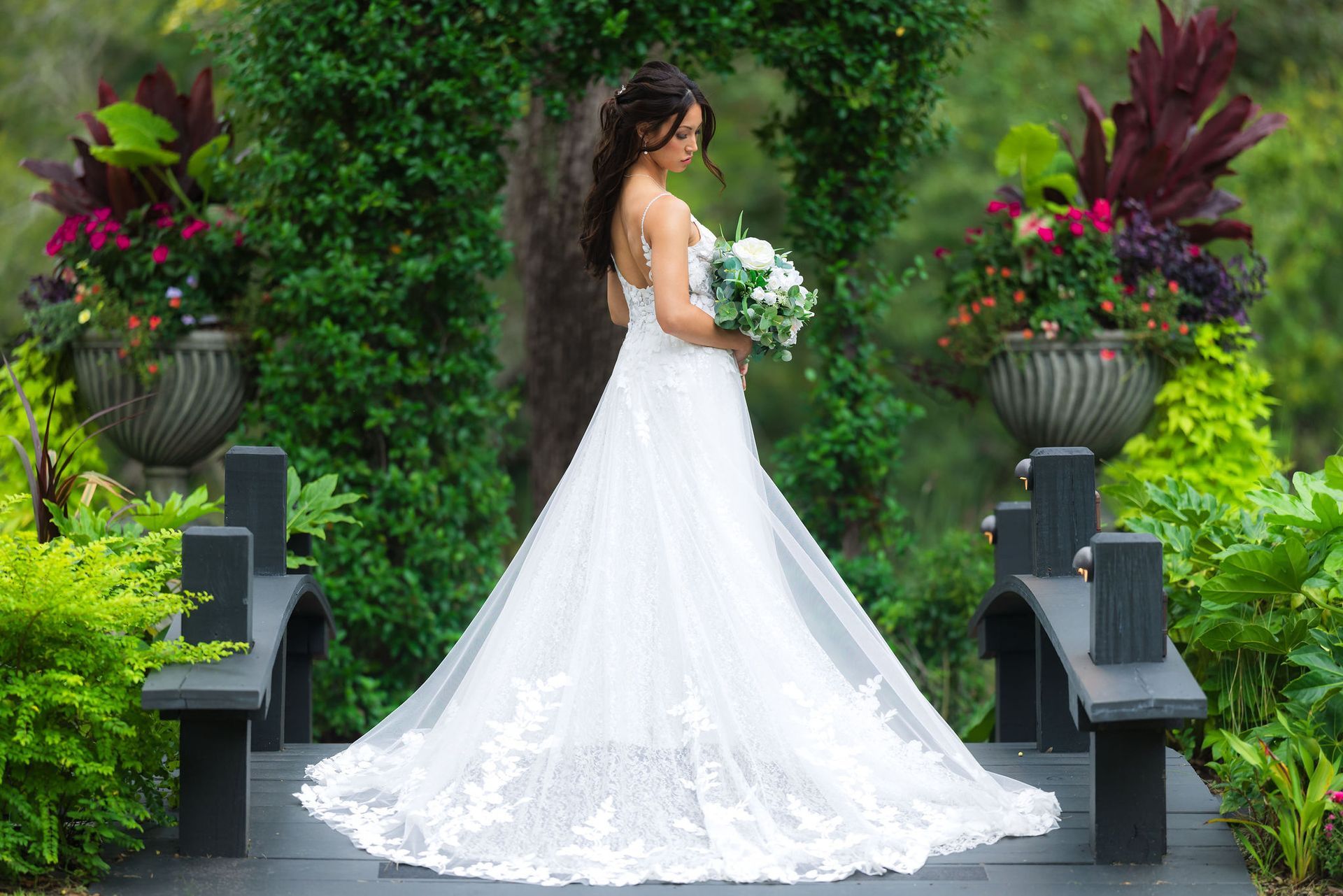 Bride in a white gown holding a bouquet on a garden path, seen from behind, with lush floral arrangements.