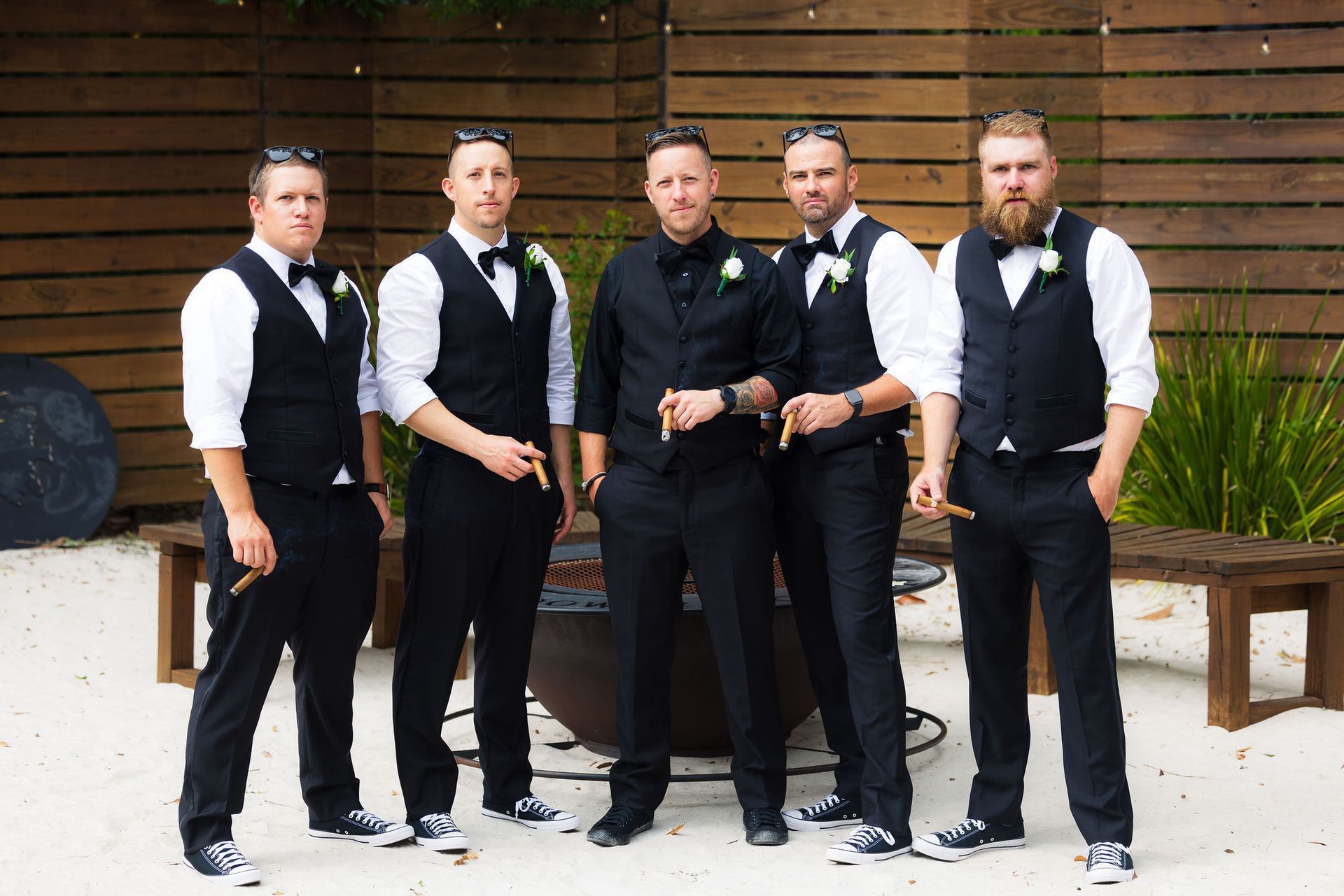 Five men in black vests and white shirts standing outdoors against a wooden wall.