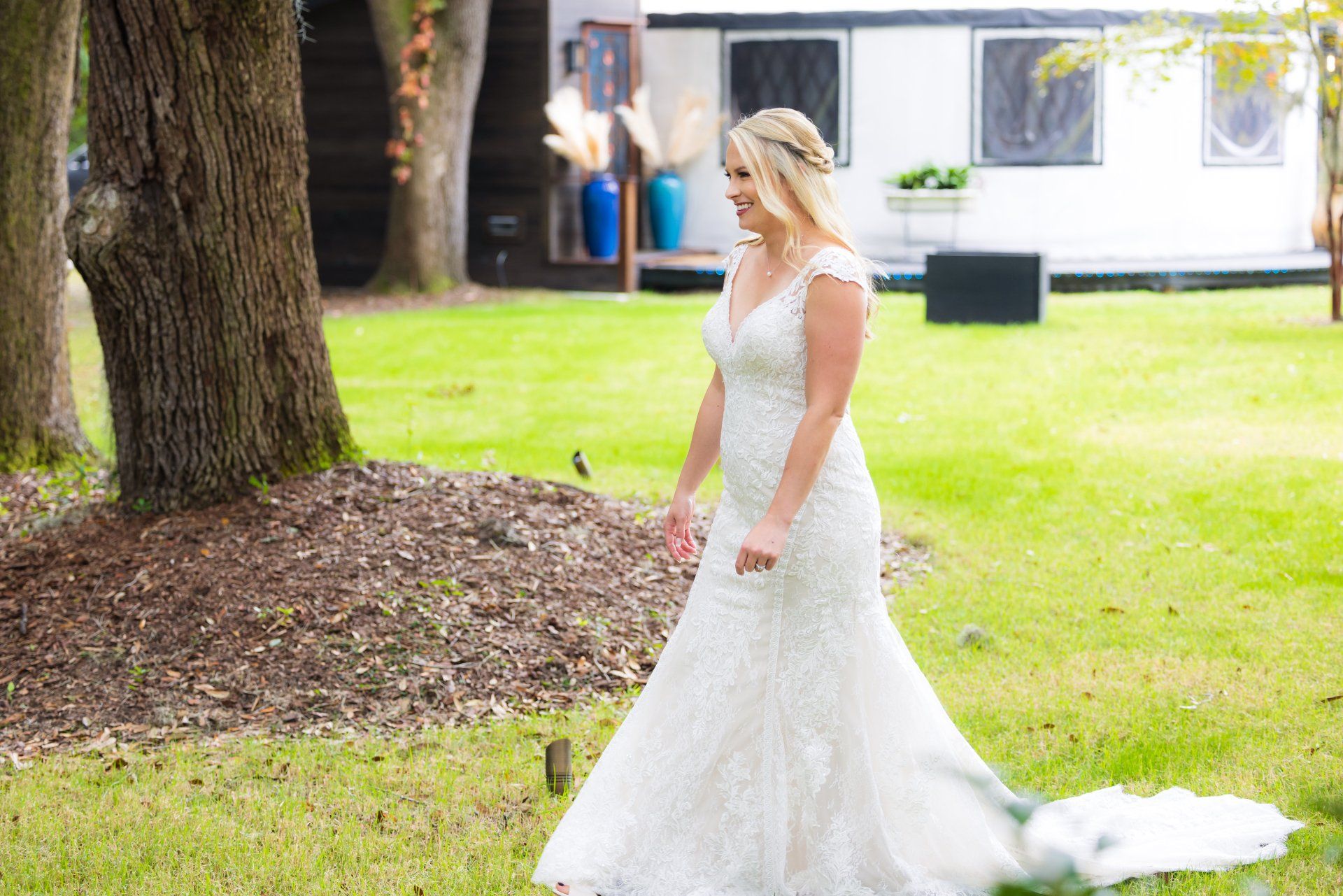 Bride in a white lace wedding dress standing on a lawn outdoors near trees and a building