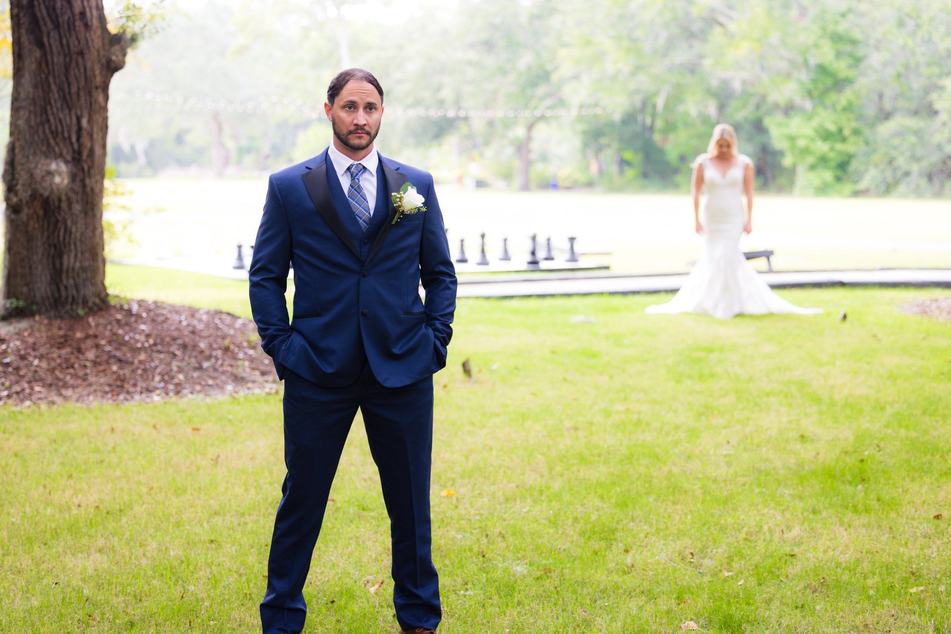 Man in a navy suit stands on grass outdoors, with a bride in a white dress near a lake in the background