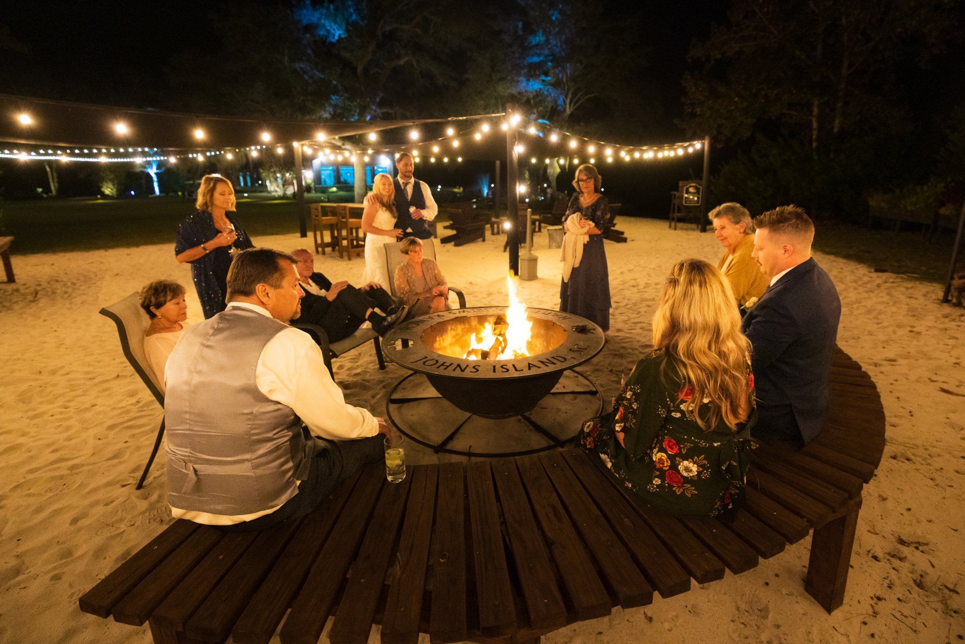 People seated around a fire pit on a sandy beach at night, with string lights overhead.