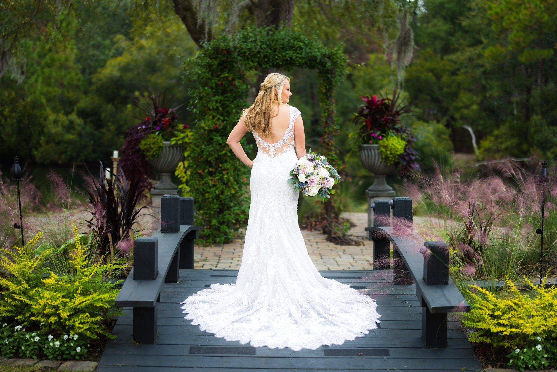 Bride in a white gown holding a bouquet on a garden walkway surrounded by greenery and flowers