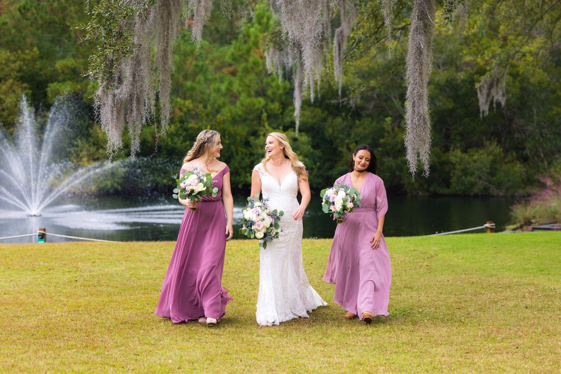 Three women in formal dresses holding bouquets on a lawn by a fountain and lake, with hanging trees behind them