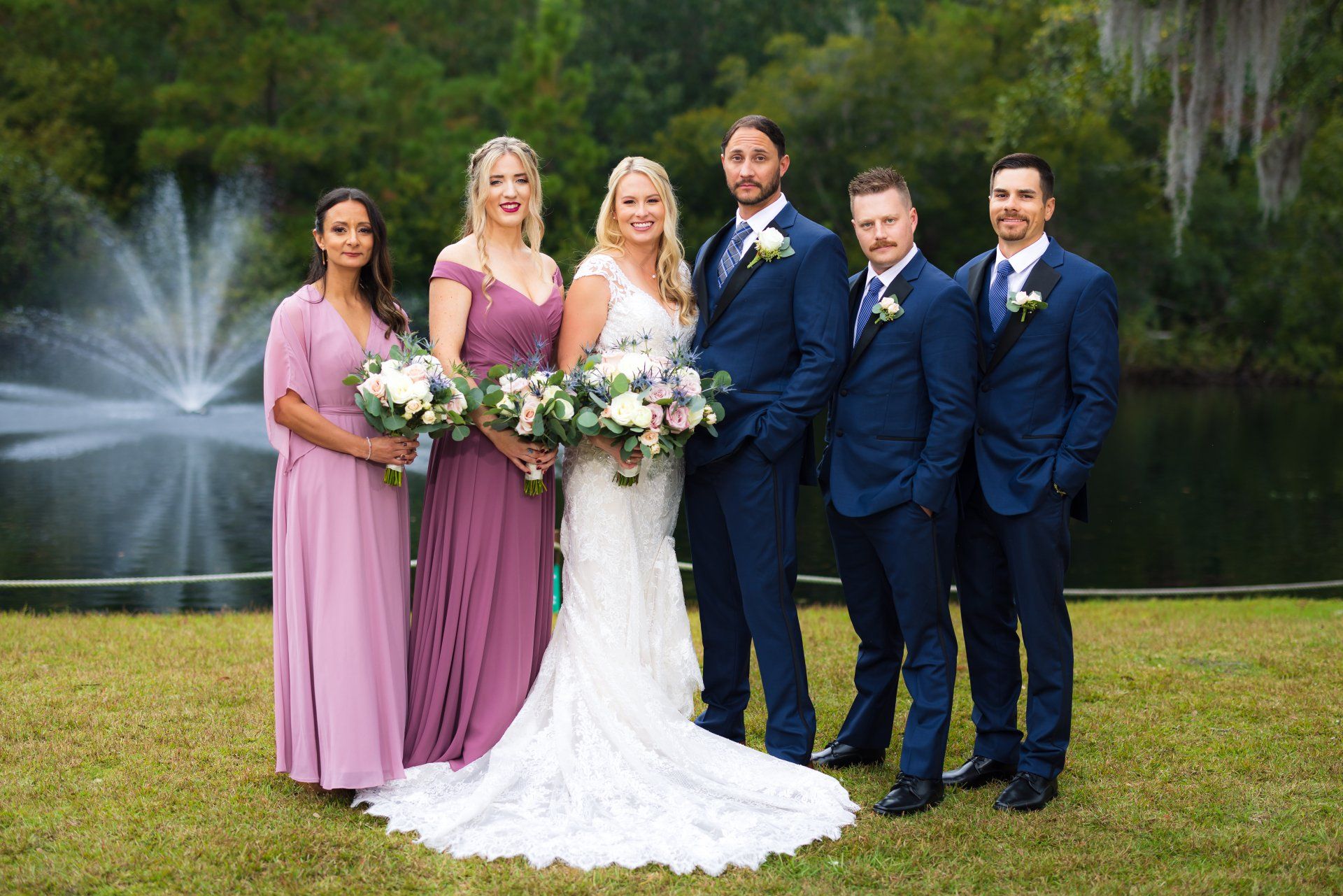 Wedding party posing outdoors by a fountain, bridesmaids in pink and the bride in a white gown, groomsmen in navy suits