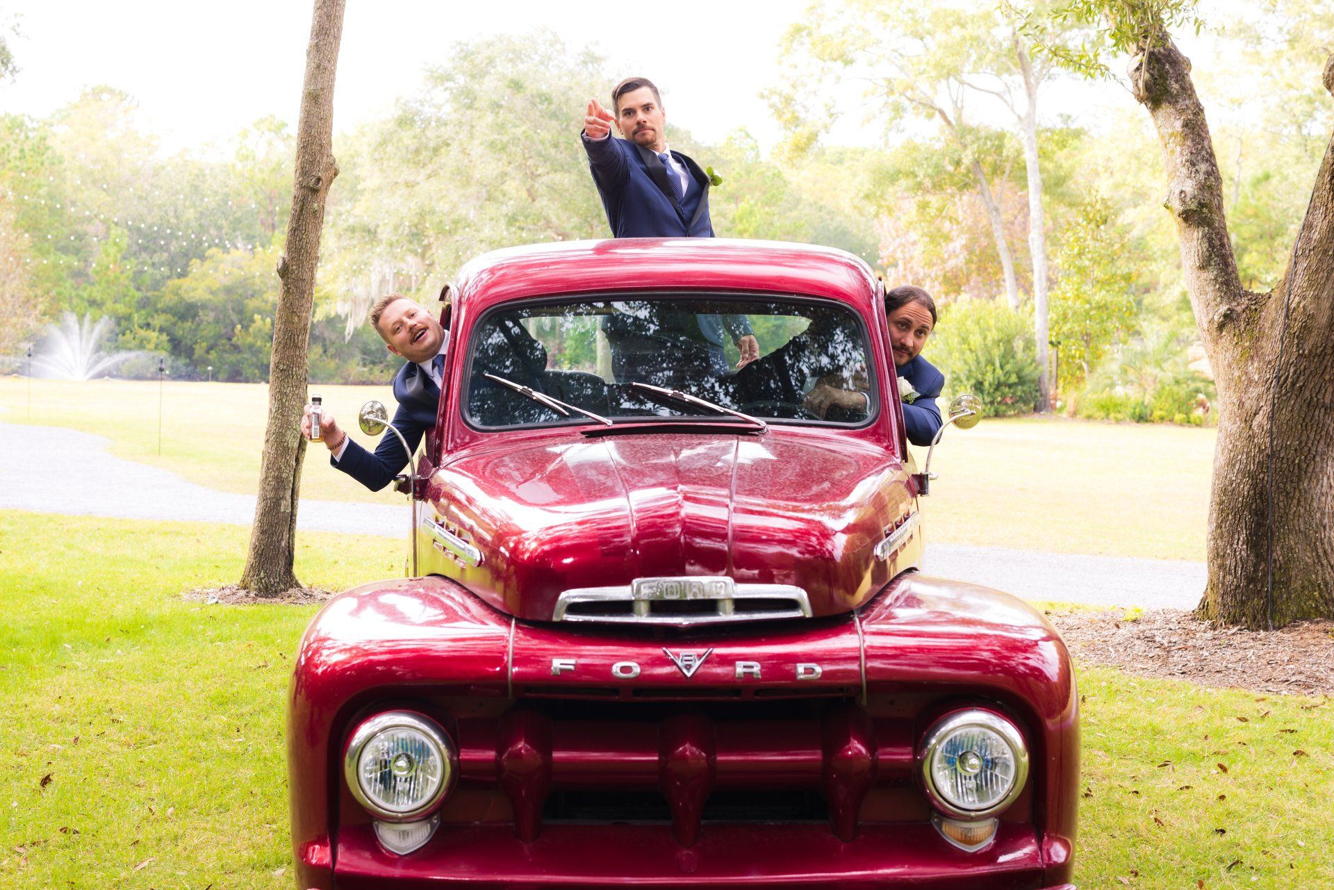 Red vintage truck with three people posing around it on a tree-lined road in a park