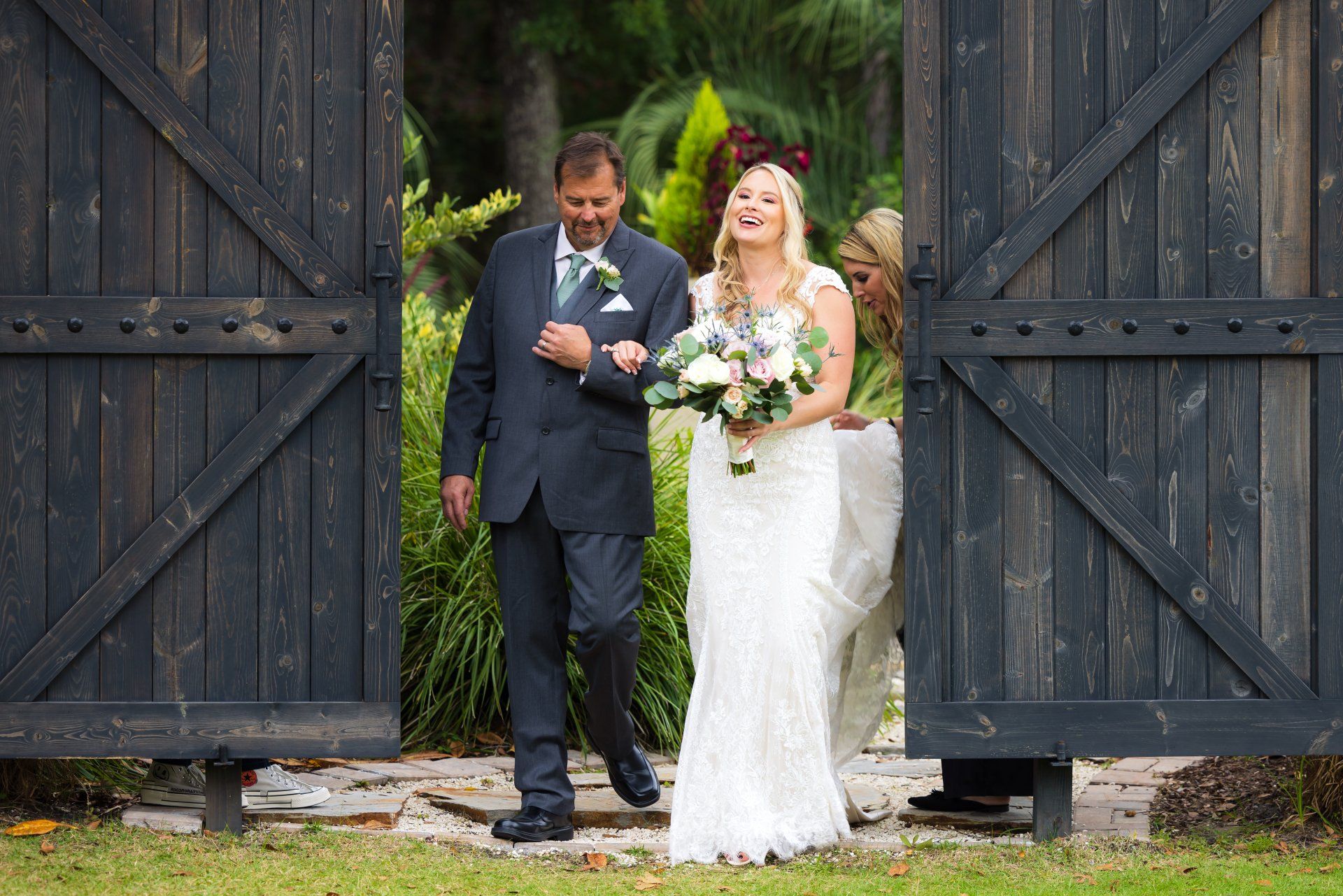 Wedding couple exiting a rustic gate, bride in white gown and groom in dark suit holding bouquets.