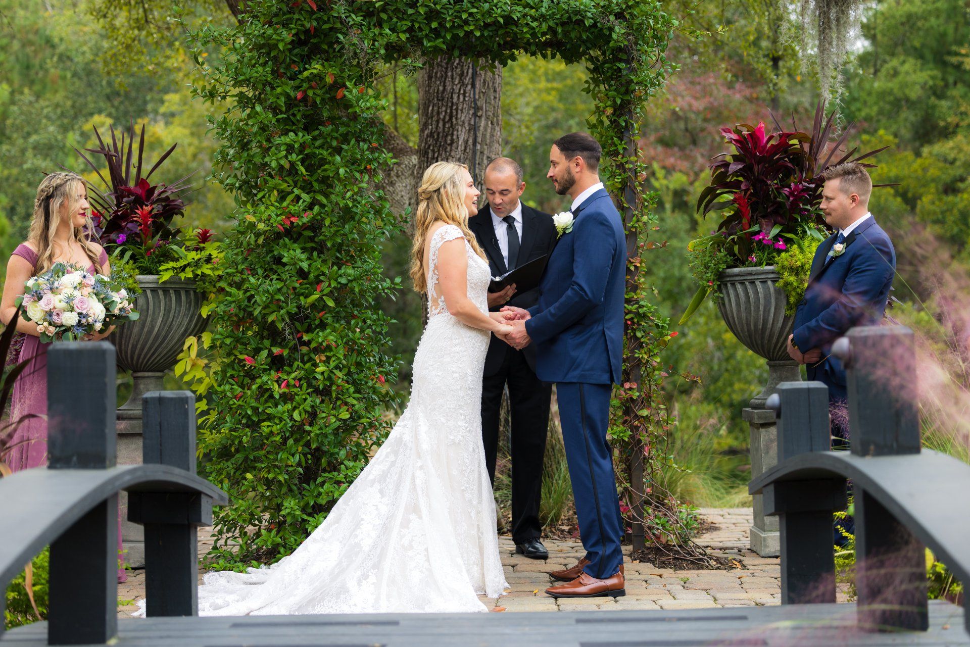 Bride and groom exchange vows outdoors on a garden bridge, with an officiant between them.