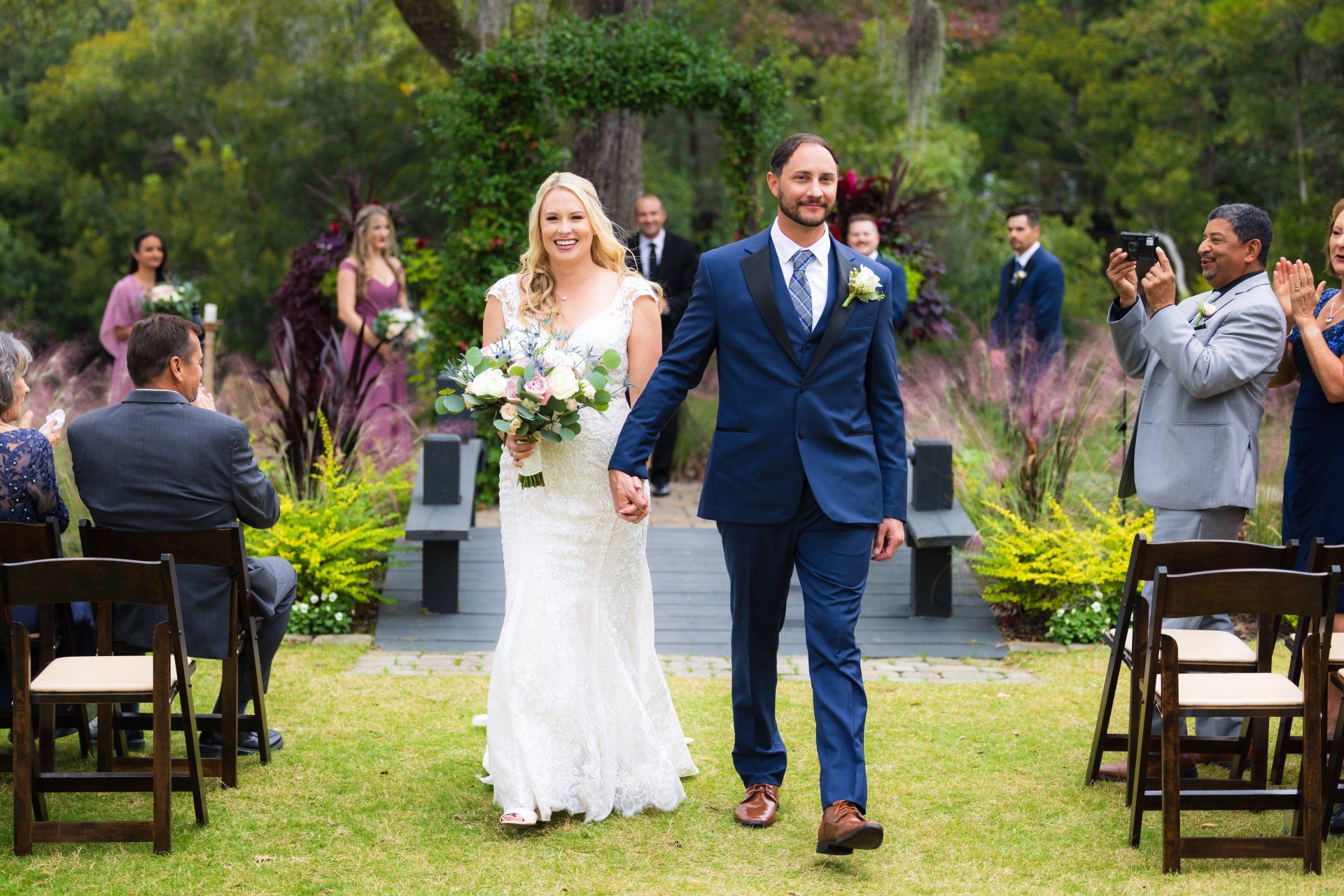 Bride and groom walk down a garden aisle after their wedding ceremony, guests applauding nearby.