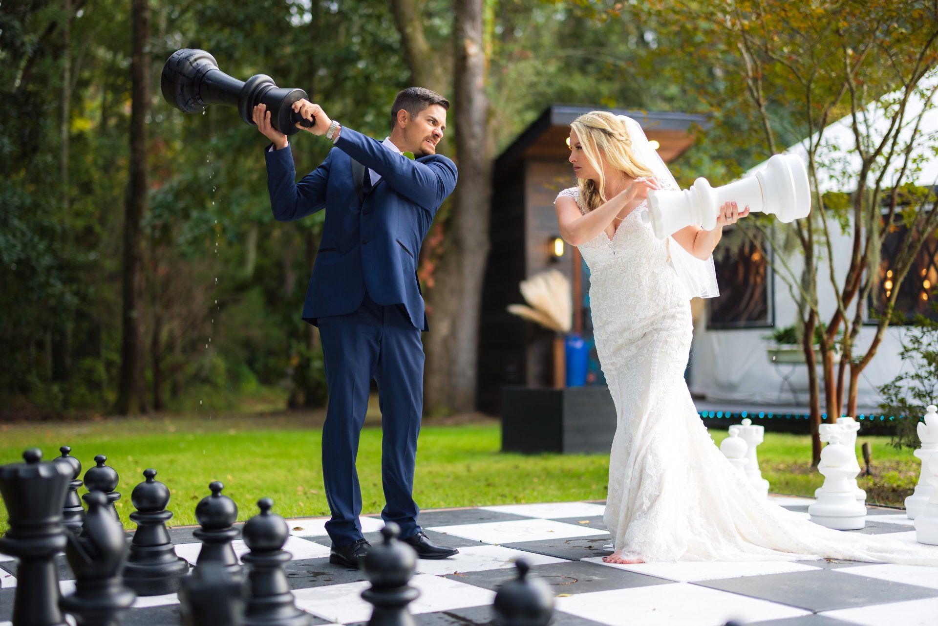 Bride and photographer posing on a lawn beside oversized chess pieces in front of a house