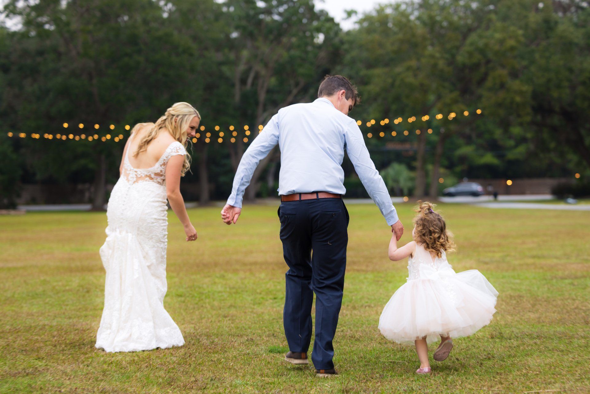 Bride, groom, and child holding hands while walking across a lawn at dusk with string lights in the background