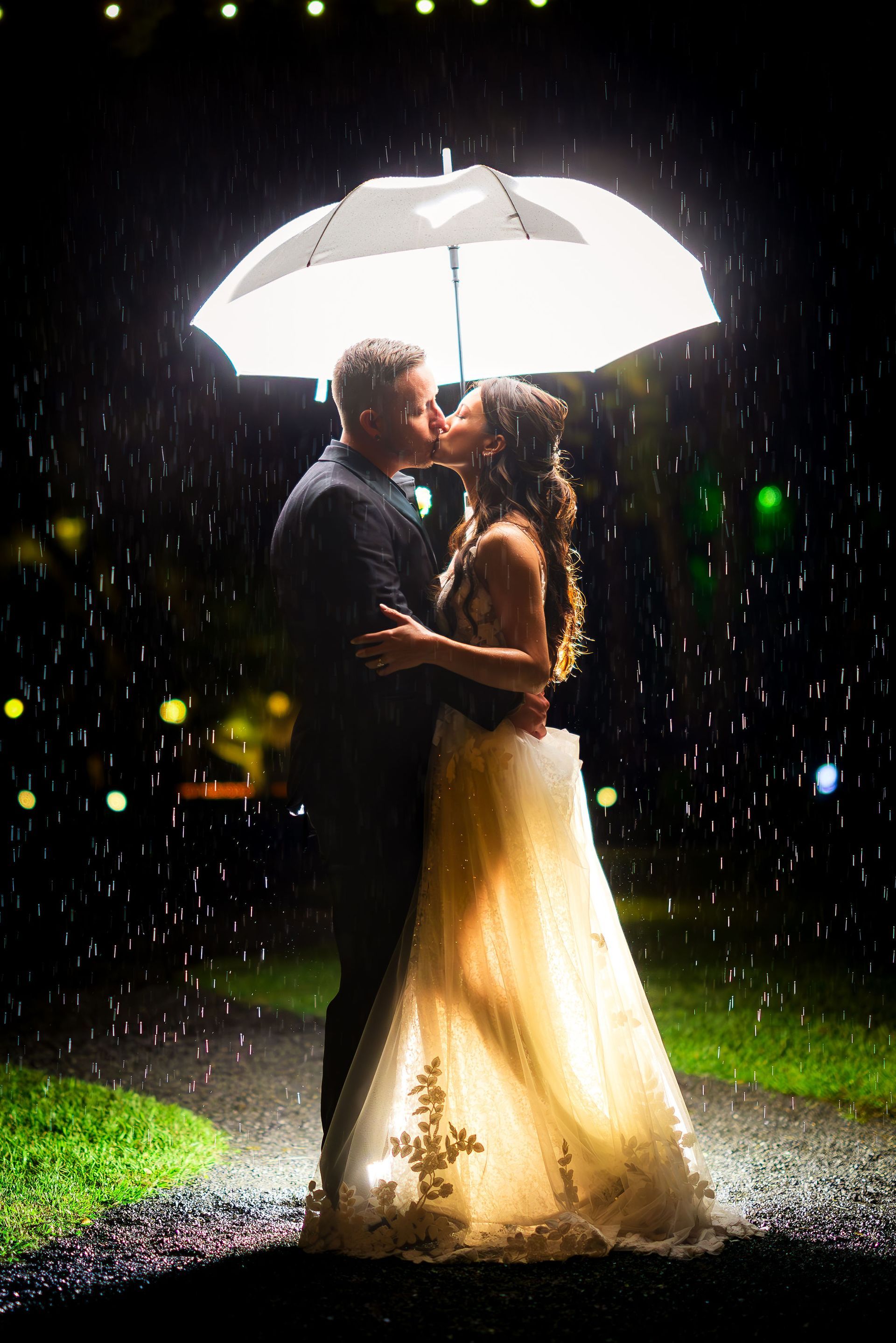 Couple kissing under a white umbrella in the rain at night, with glowing lights behind them.