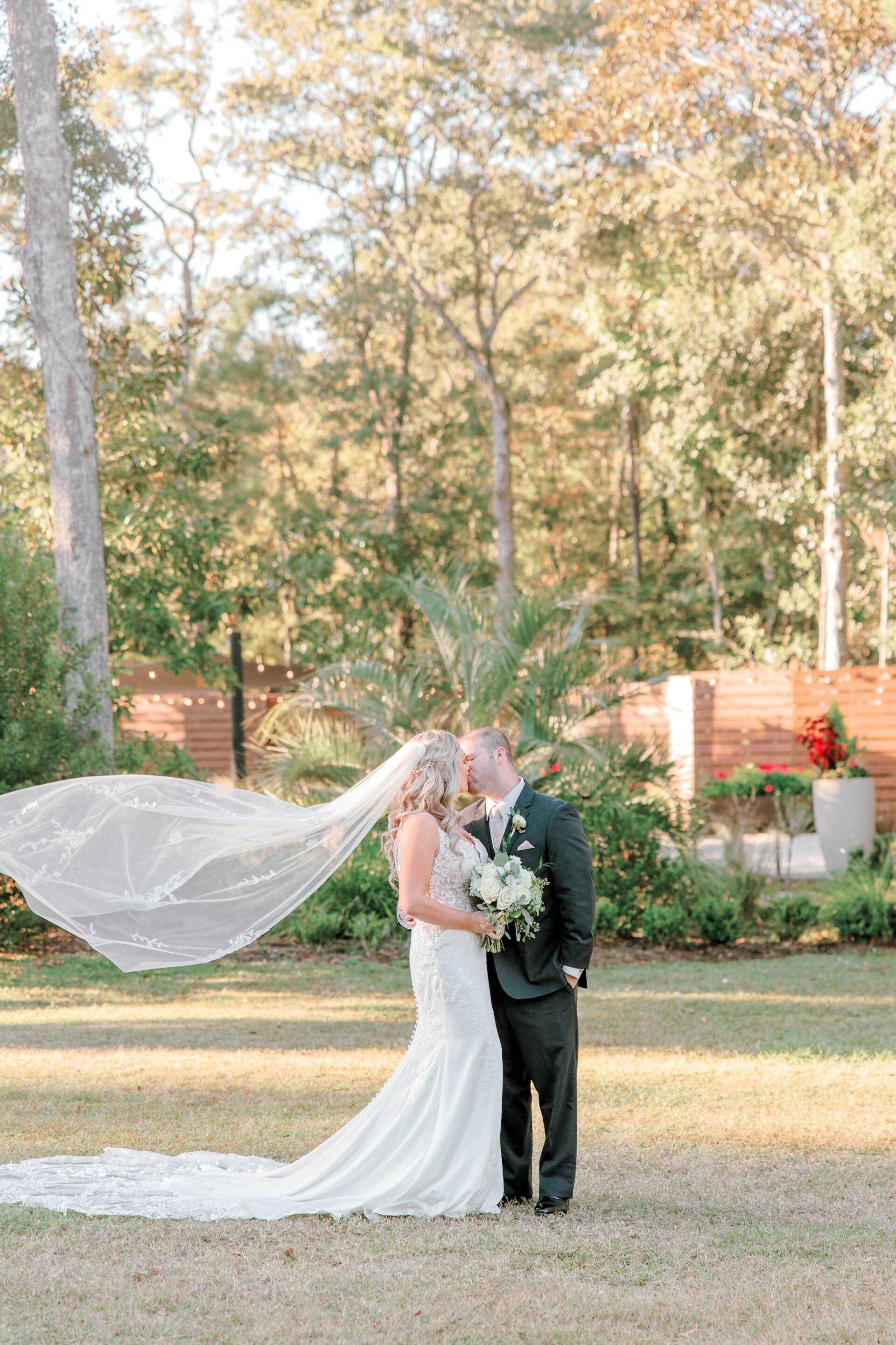 Bride and groom in a garden, with the bride’s long veil blowing in the wind.