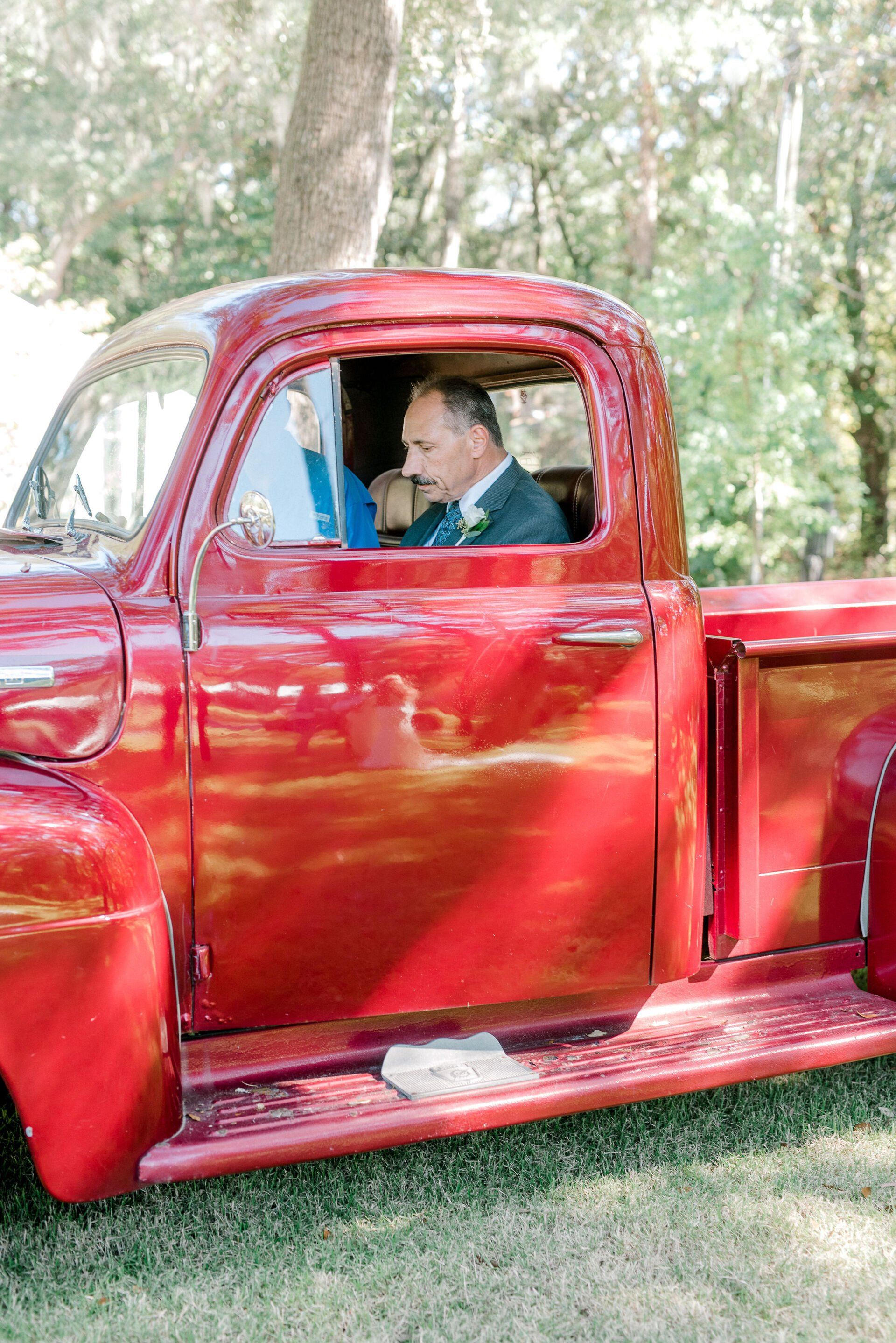 Man sitting in a shiny red vintage truck outdoors on grass, with trees in the background