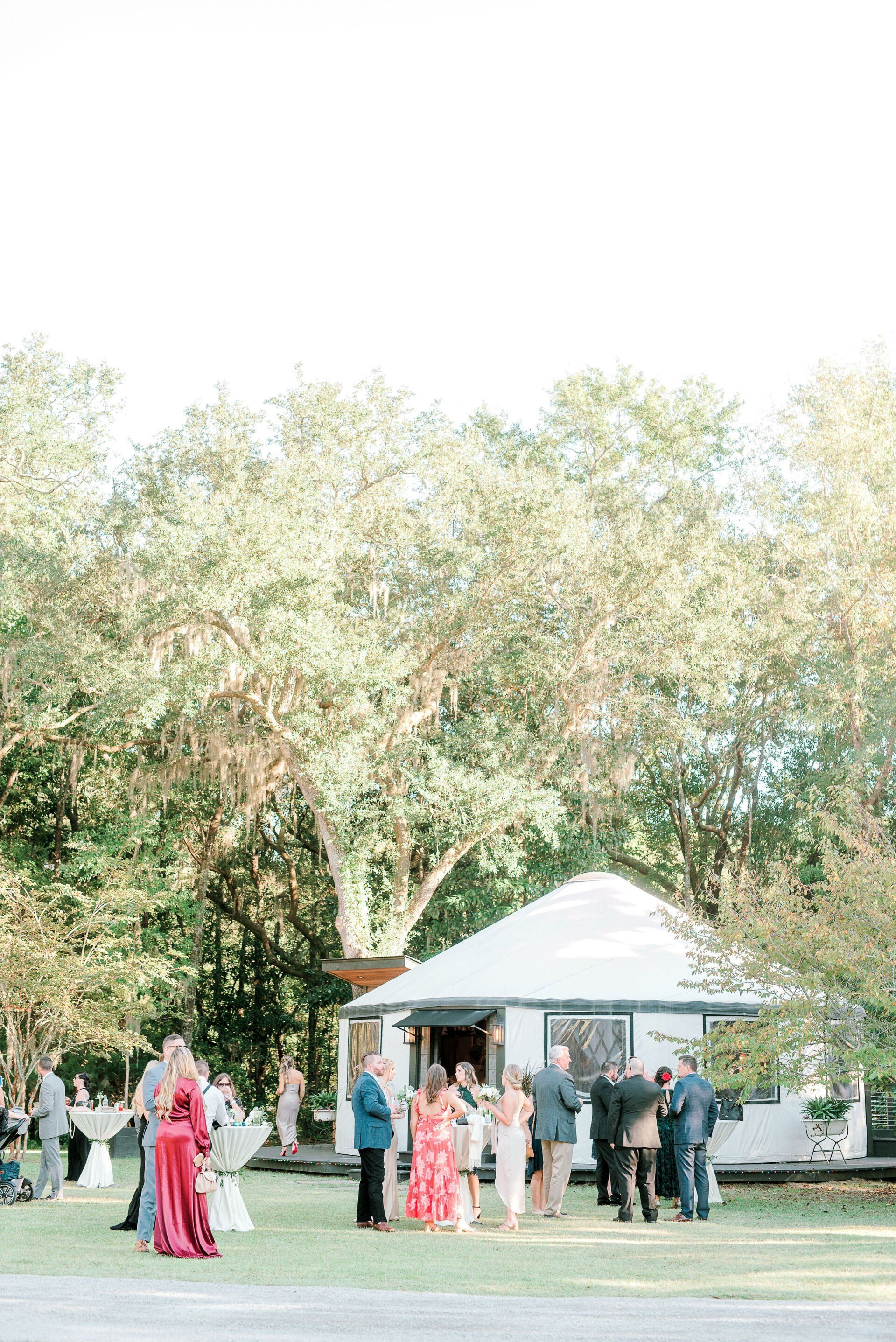 People gathered near a white tent outdoors under tall trees, some in formal attire.