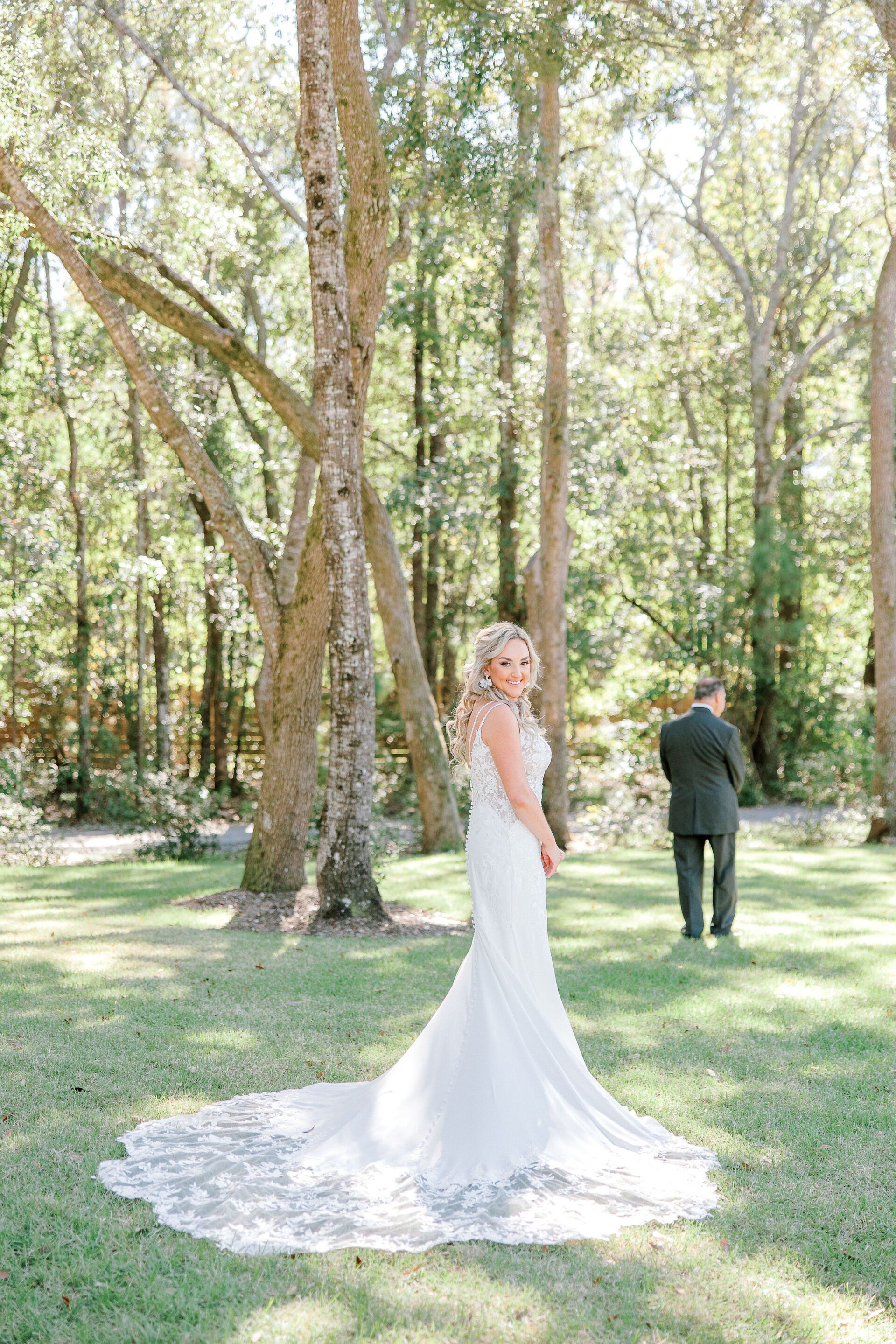Bride in a white gown standing in a sunlit wooded garden, with a groom in the background.