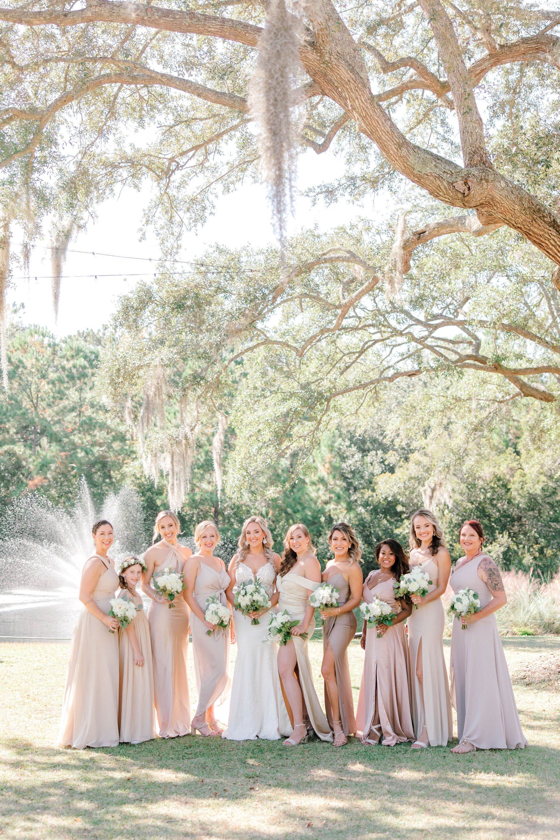 Bridal party in blush dresses holding bouquets in a sunlit garden under trees