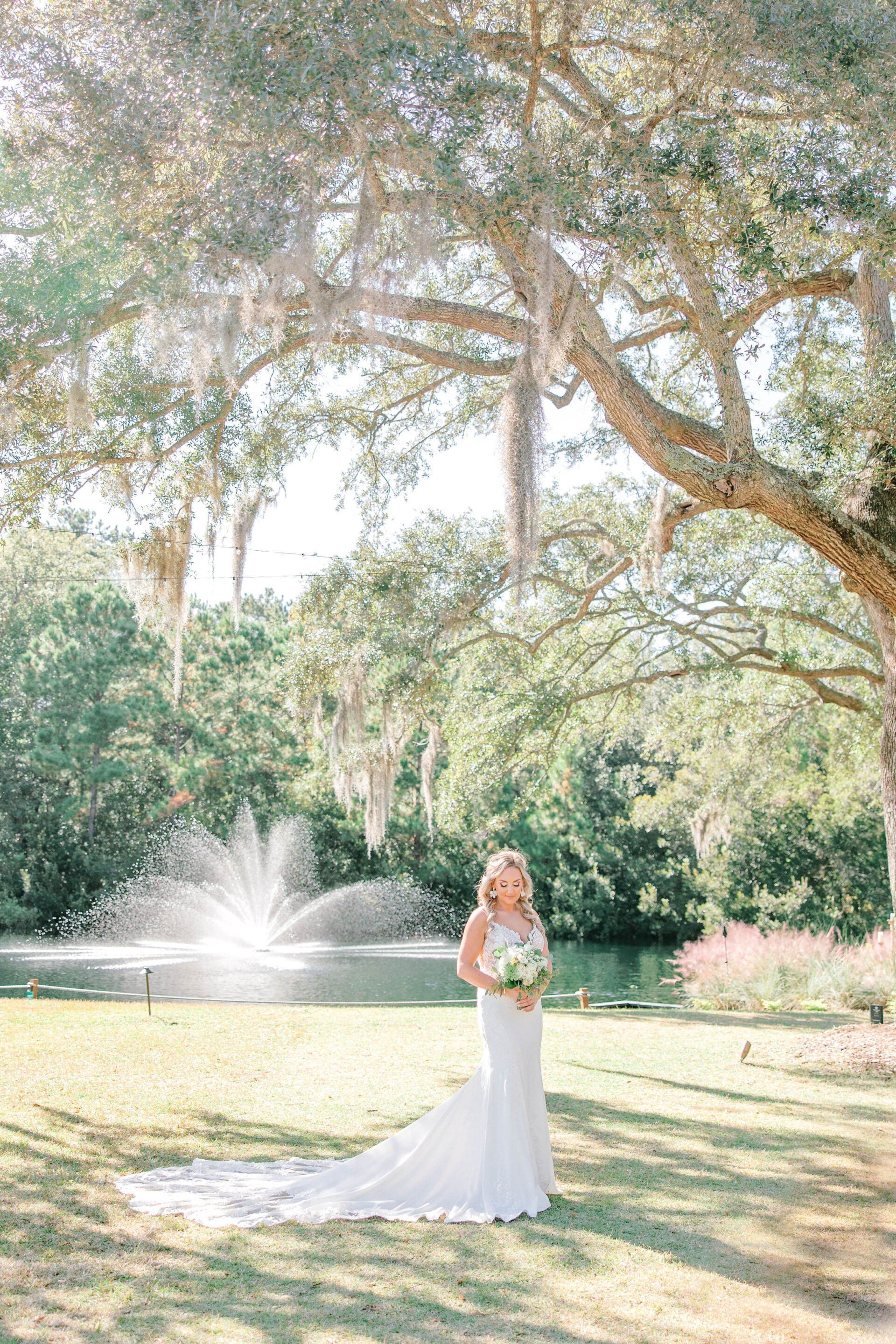 Bride in a white gown holding flowers in a sunlit garden with a fountain and trees.