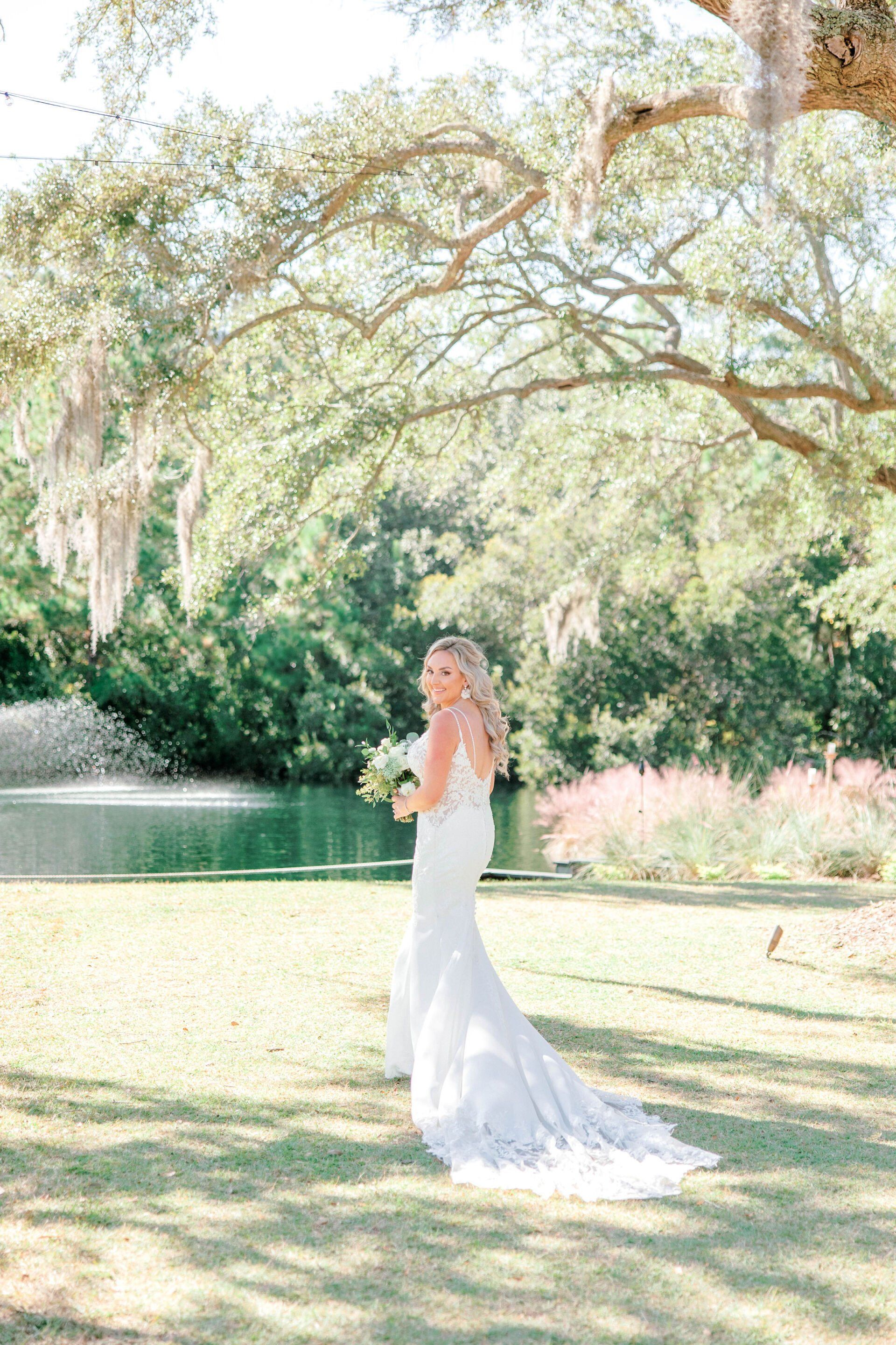 Woman in a white gown standing on a sunlit lawn beneath sprawling trees near a pond.