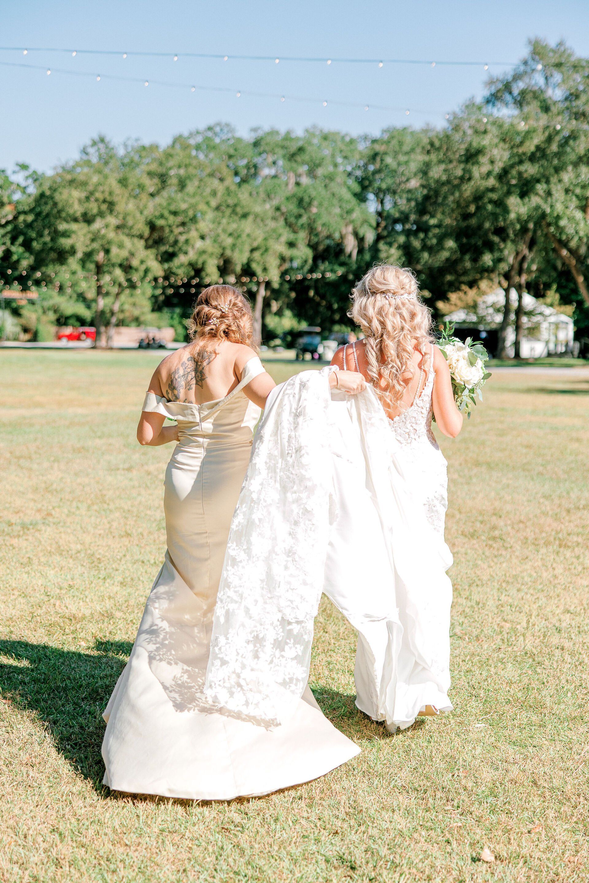Two brides in white wedding dresses walking across a sunny lawn, seen from behind