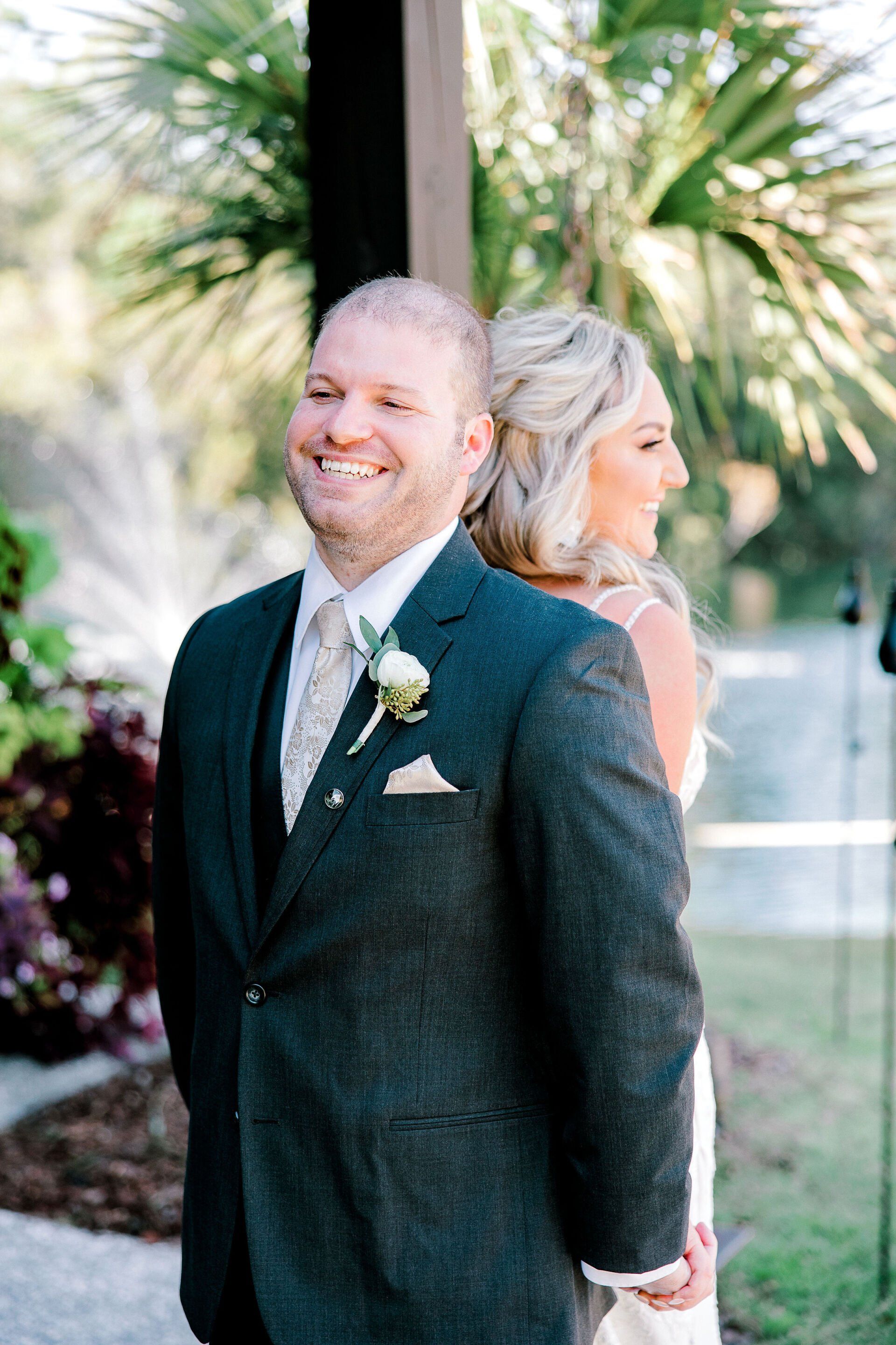 Smiling couple standing back to back outdoors, with palm trees and greenery behind them.