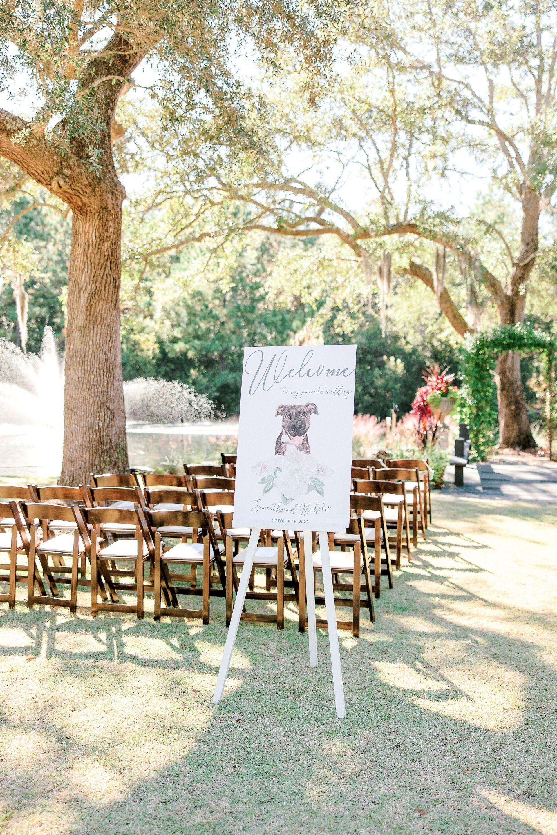 Outdoor wedding ceremony setup with wooden chairs, an easel sign, and sunlit trees lining the aisle