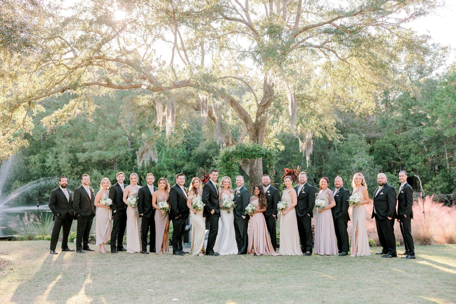 Wedding party posing outdoors in a garden, with trees and fountain in the background.