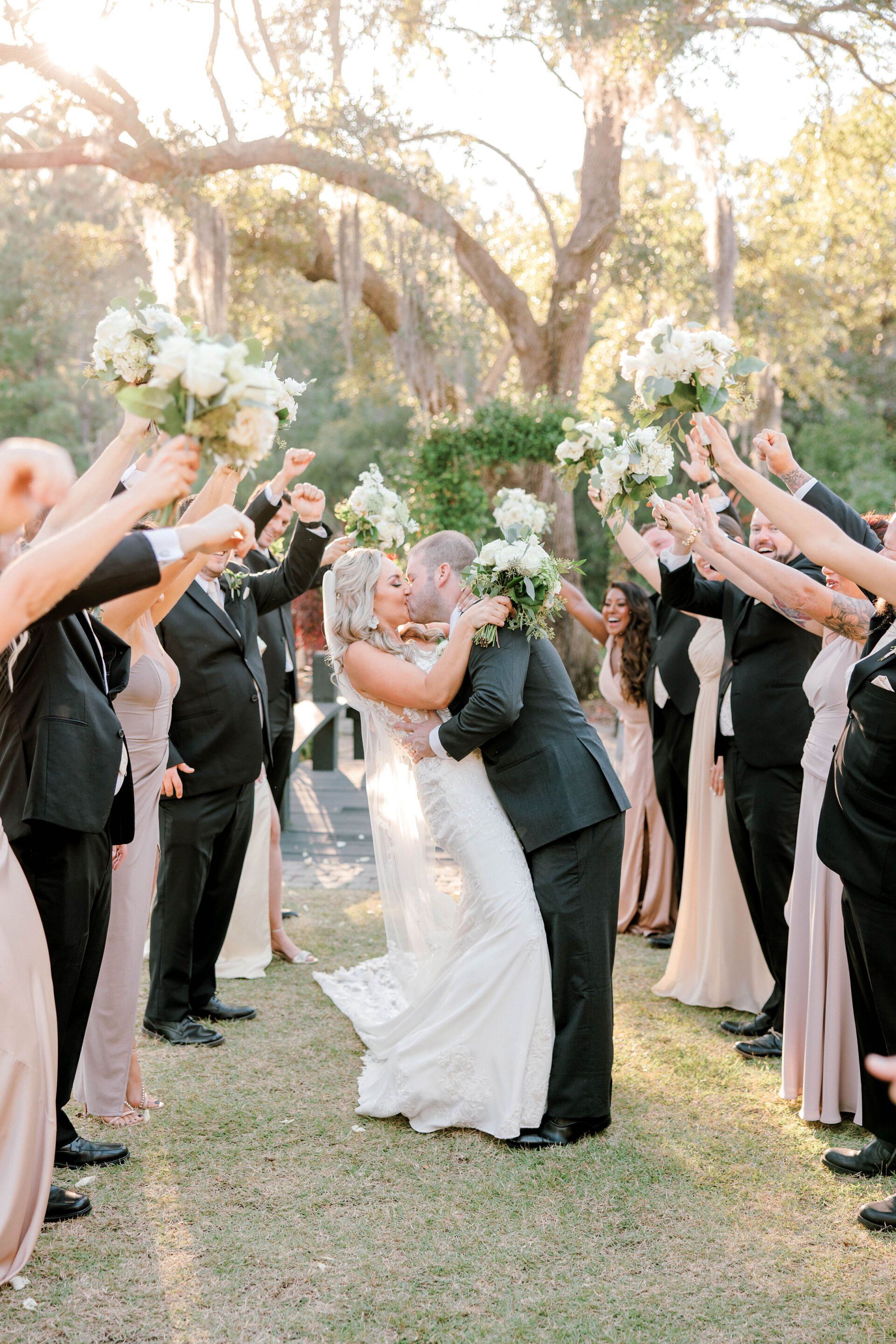 Wedding couple kissing under a flower arch with guests raising bouquets outdoors
