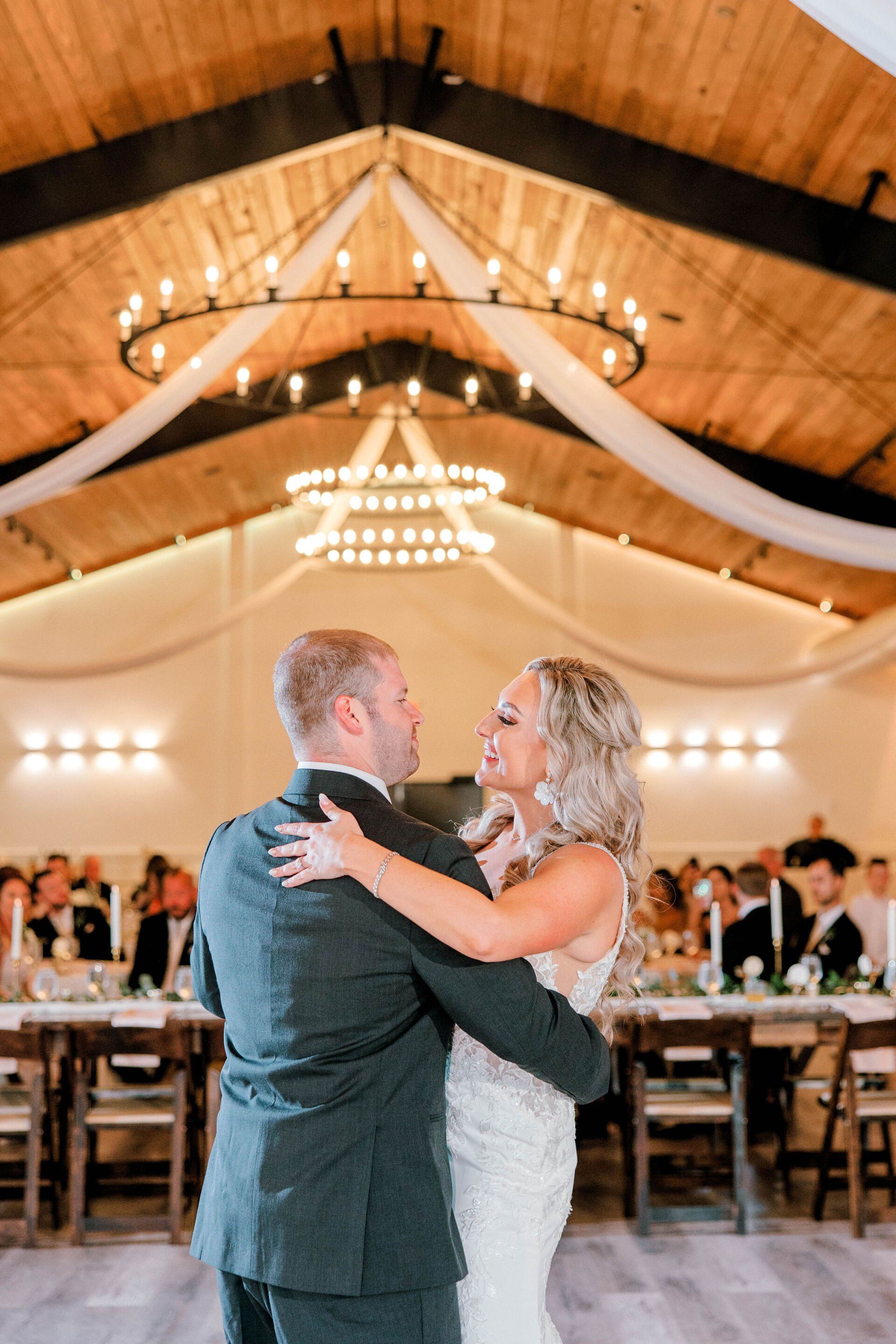 Bride and groom dancing in a warmly lit reception hall with wooden beams and draped fabric