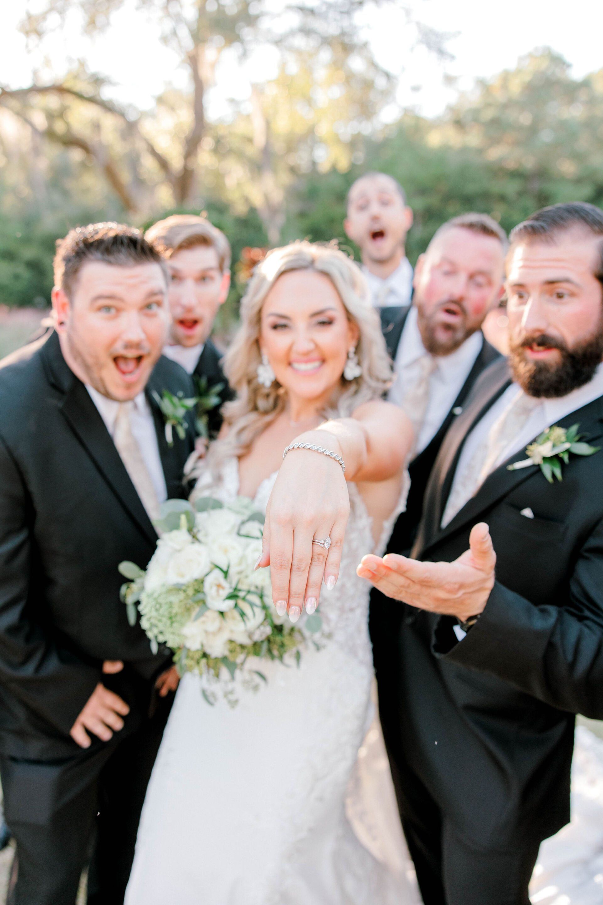 Wedding party posing outdoors, smiling and cheering around the bride in a white dress holding a bouquet.