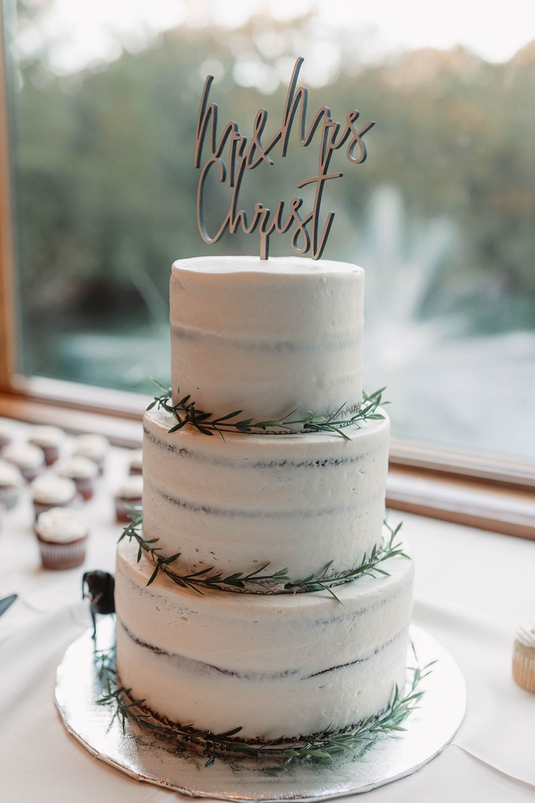 Three-tier white wedding cake with greenery accents and a handwritten topper by a window