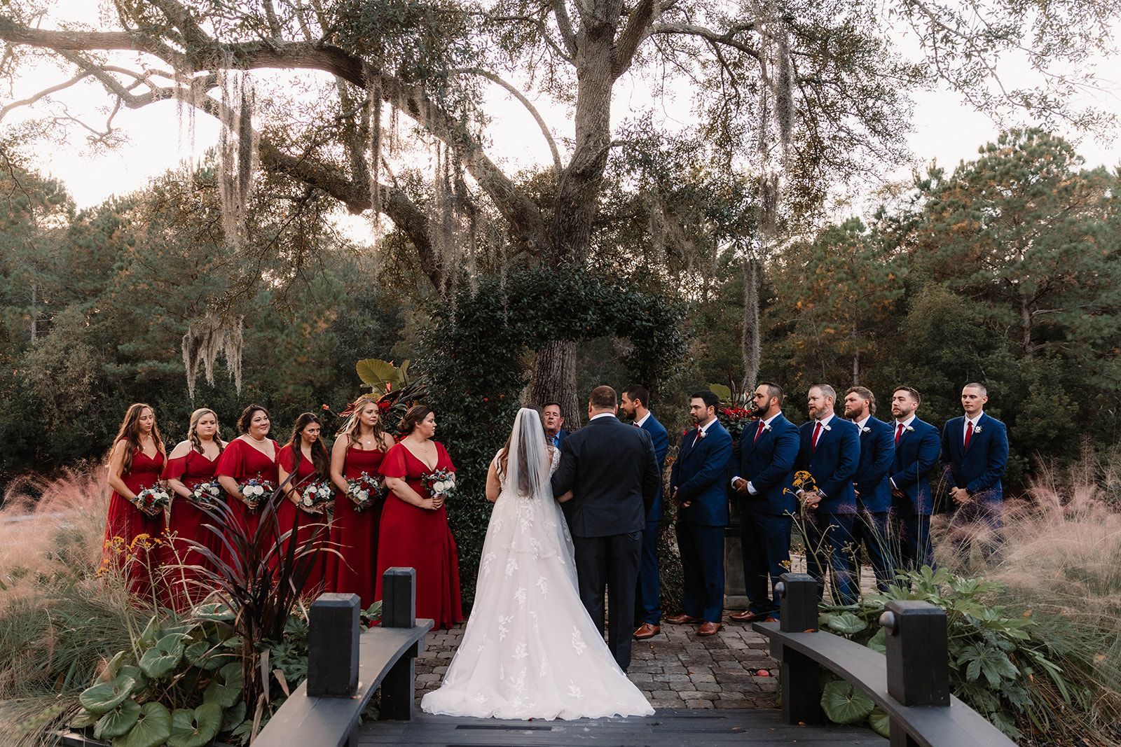 Wedding ceremony outdoors with bride in white and bridal party in red and blue standing beneath trees