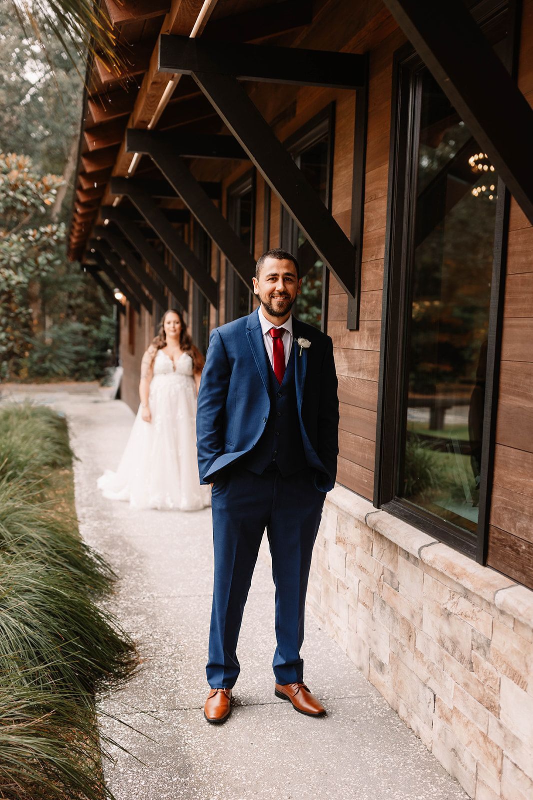 Man in blue suit stands under a wooden covered walkway; bride in white dress walks behind him.