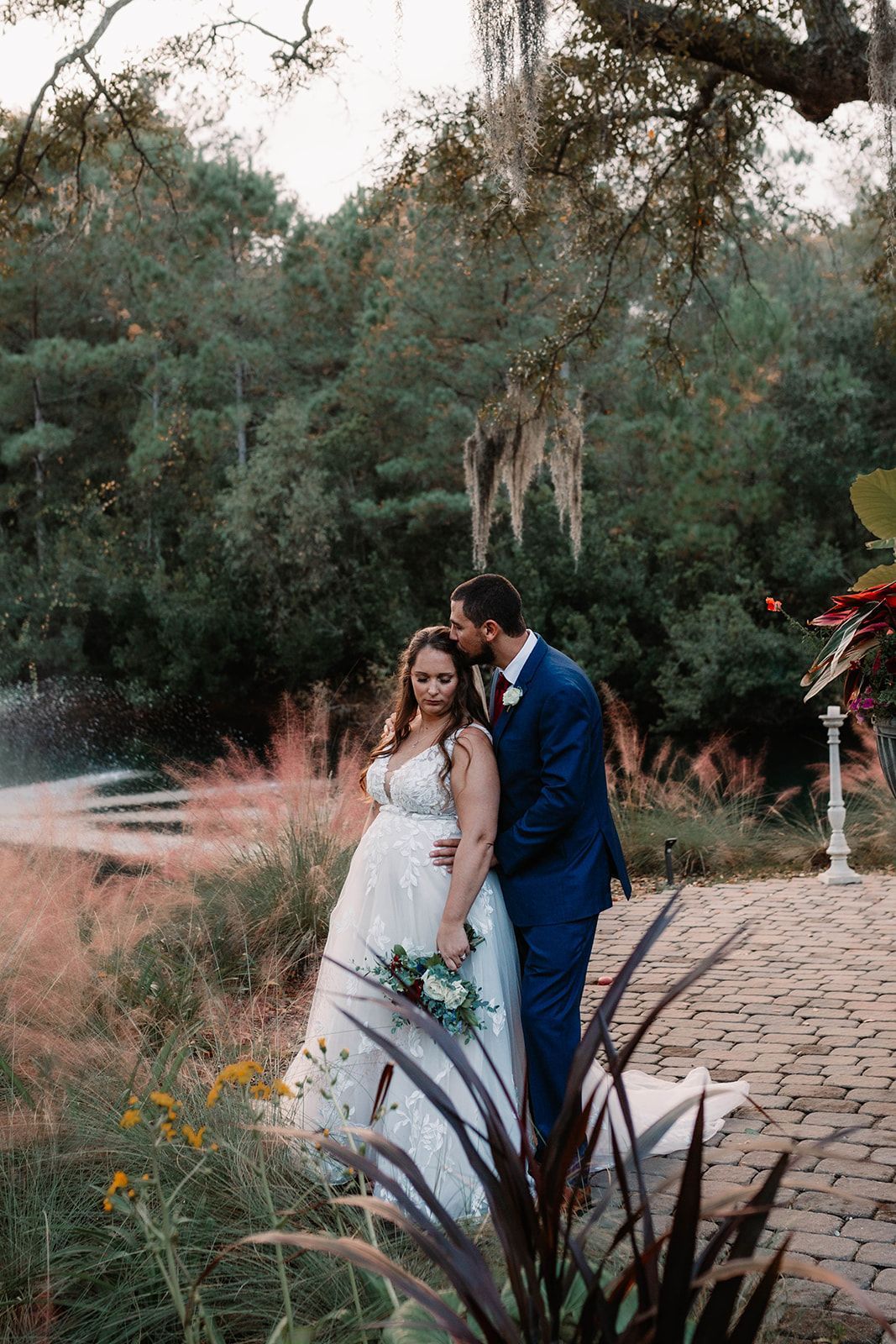 Bride and groom posing outdoors on a stone path in a garden, surrounded by greenery and tall grasses