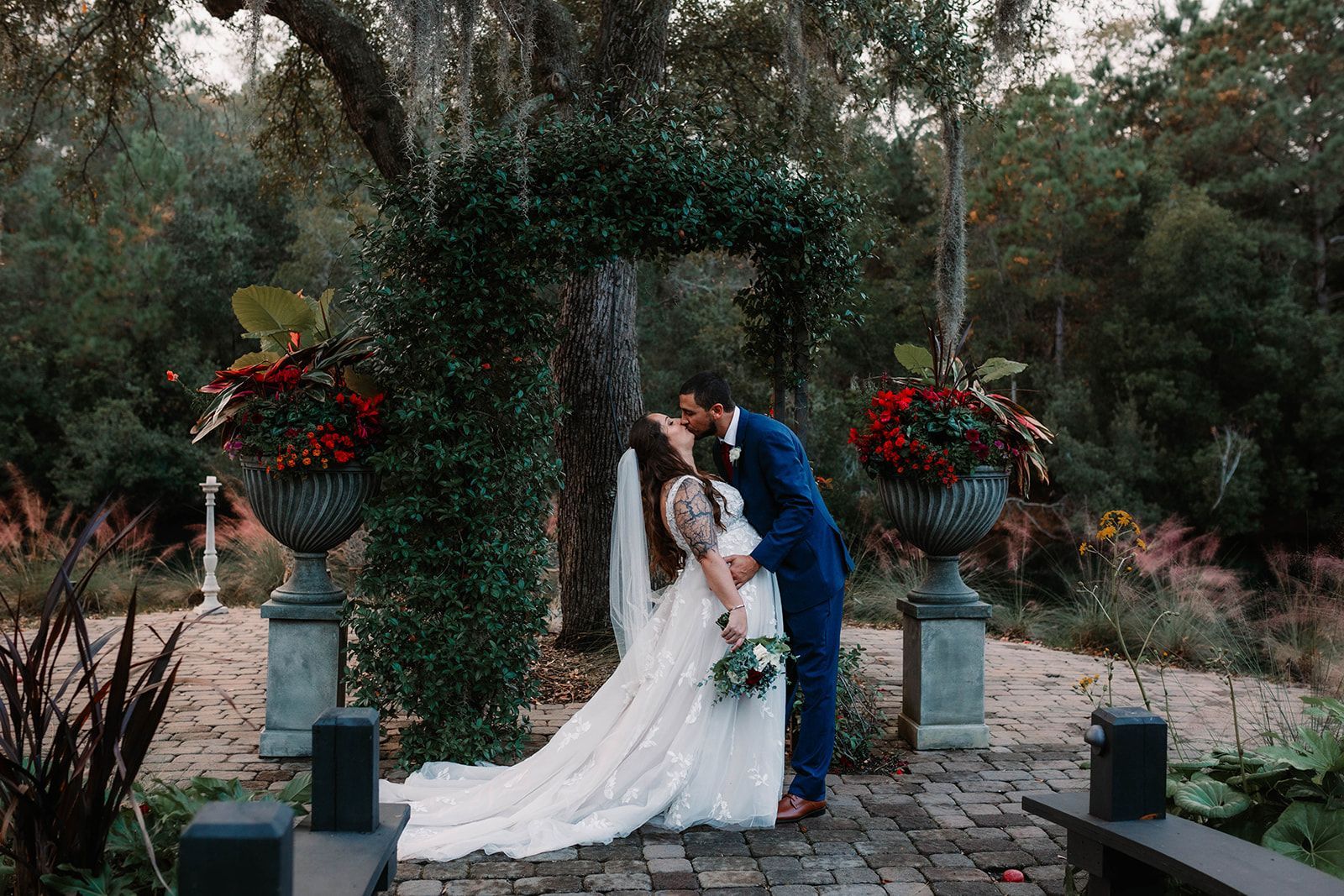 Bride and groom kissing in a garden gazebo, surrounded by red flower arrangements.