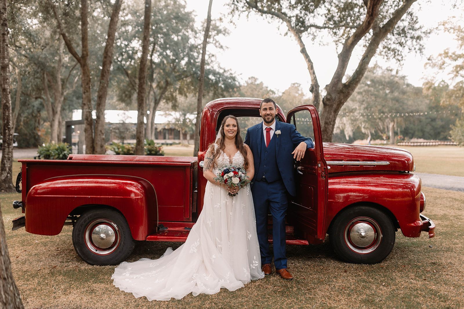 Bride and groom posing beside a red vintage pickup truck in a wooded outdoor setting