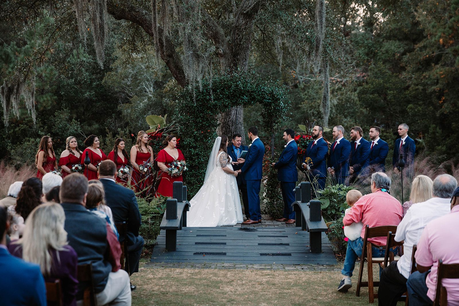 Outdoor wedding ceremony with bride and groom at altar, attendants in red and blue, seated guests watching