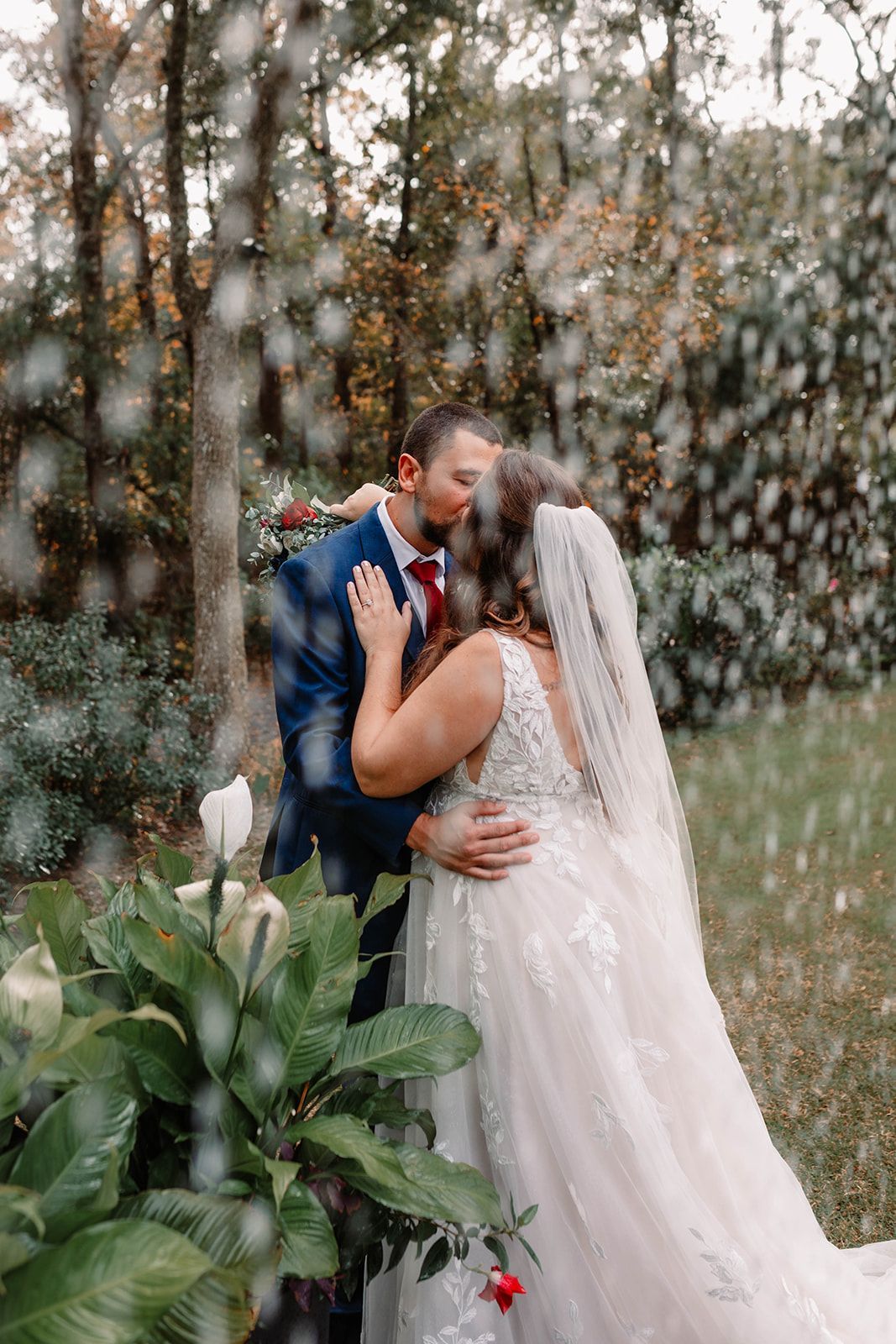 Bride and groom kiss in a wooded outdoor setting with white flowers and falling snow.