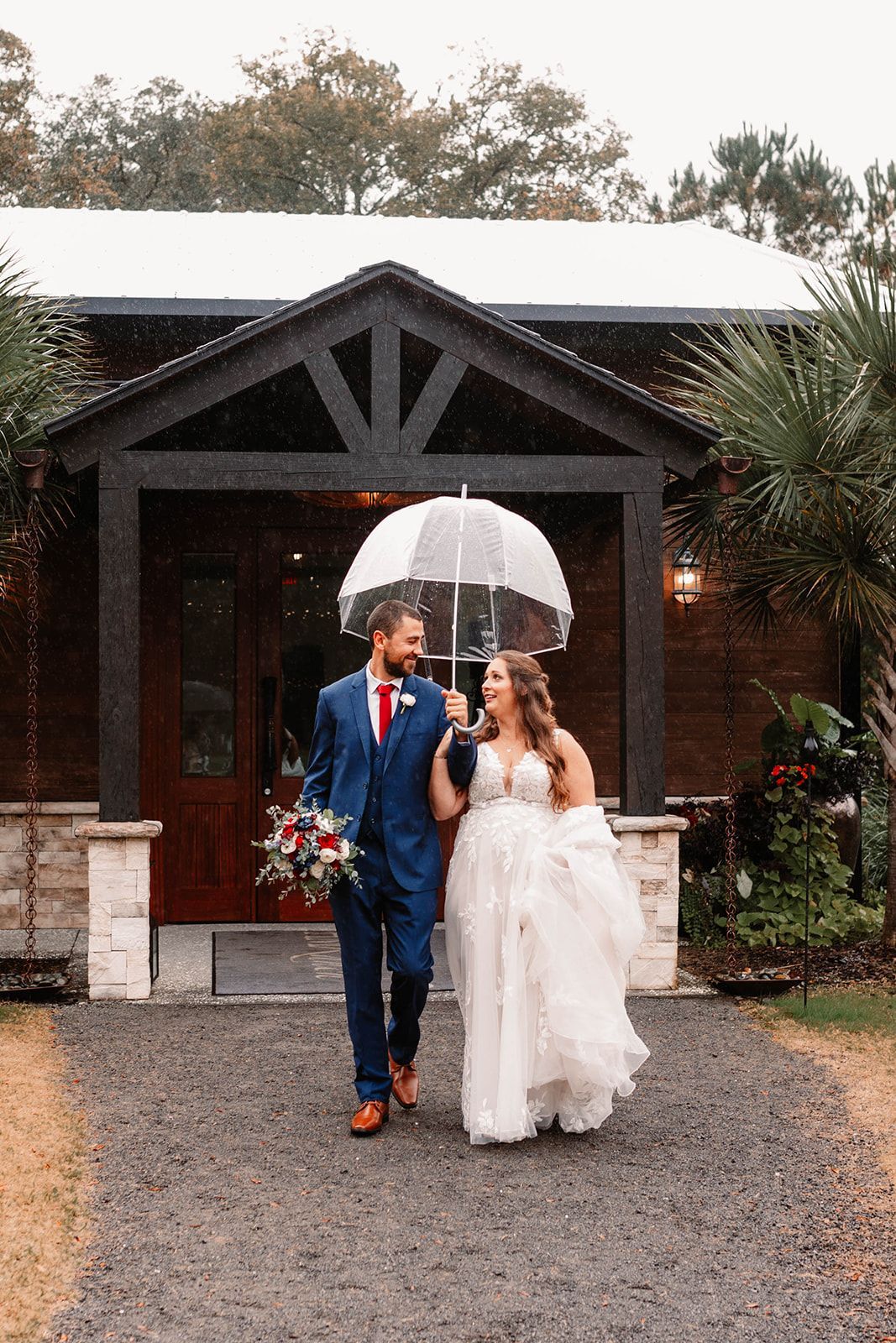 Bride and groom in wedding attire walking under a clear umbrella outside a rustic building