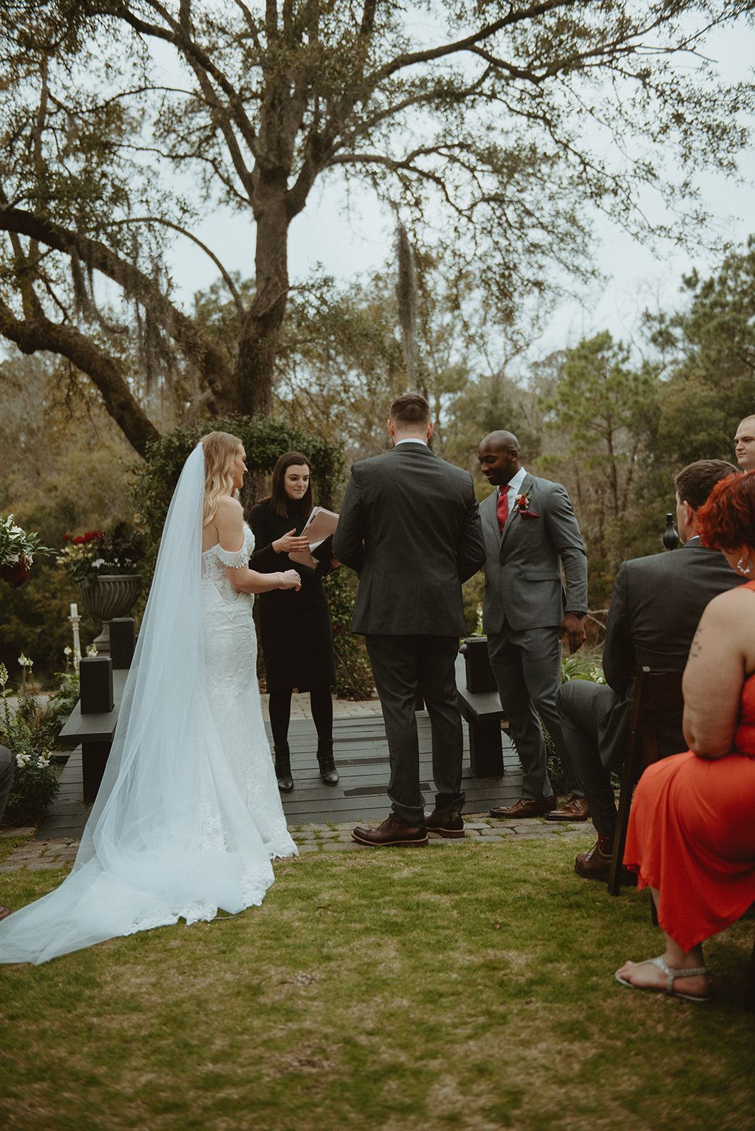 Bride and groom at an outdoor wedding ceremony under trees, with guests nearby.