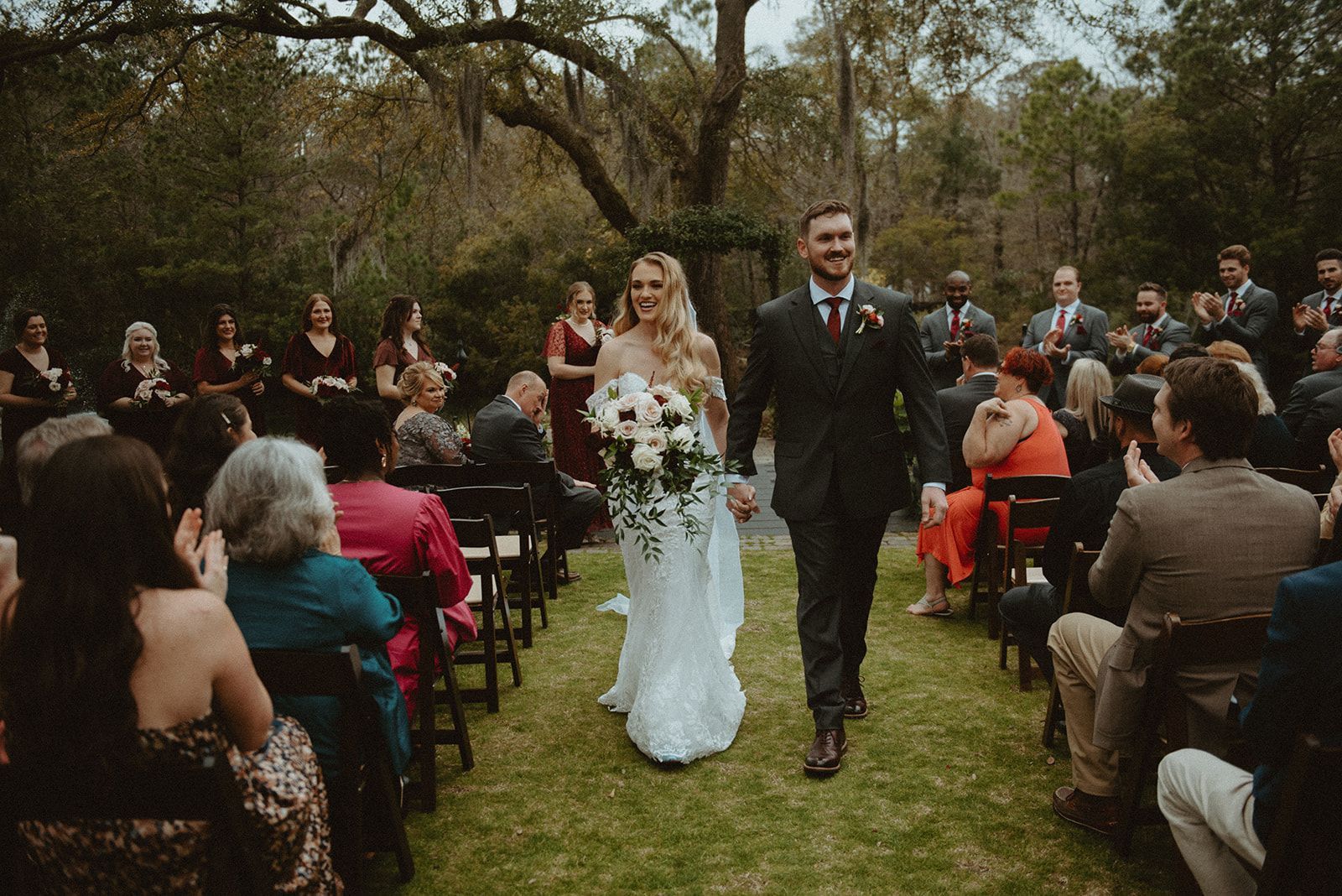 Newlyweds walking down an outdoor aisle at a tree-lined wedding ceremony, guests seated on both sides.