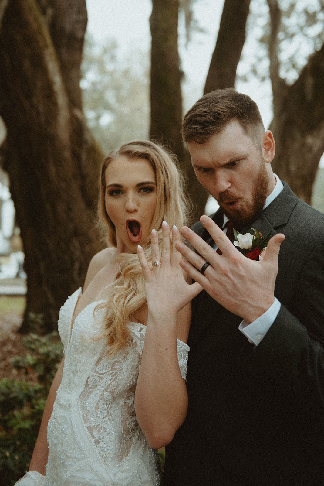 Bride and groom posing in a wooded setting, both making surprised expressions and showing their rings.