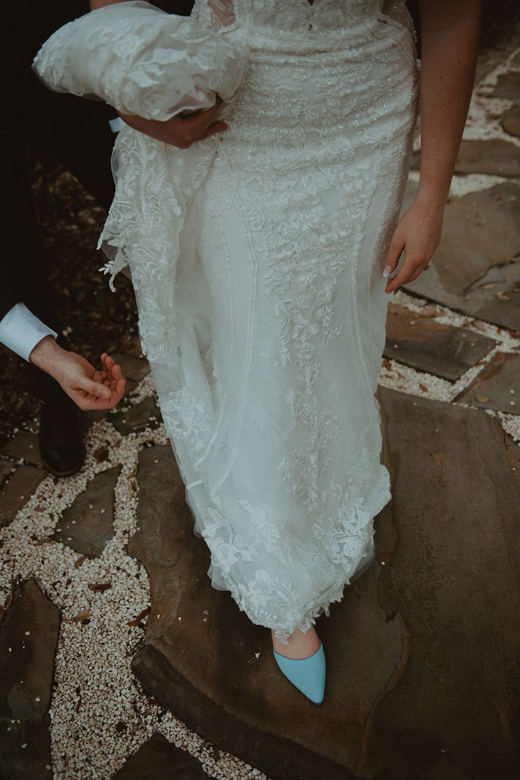 Bride in a white lace dress and blue heels walking on a stone path, holding a white bouquet.