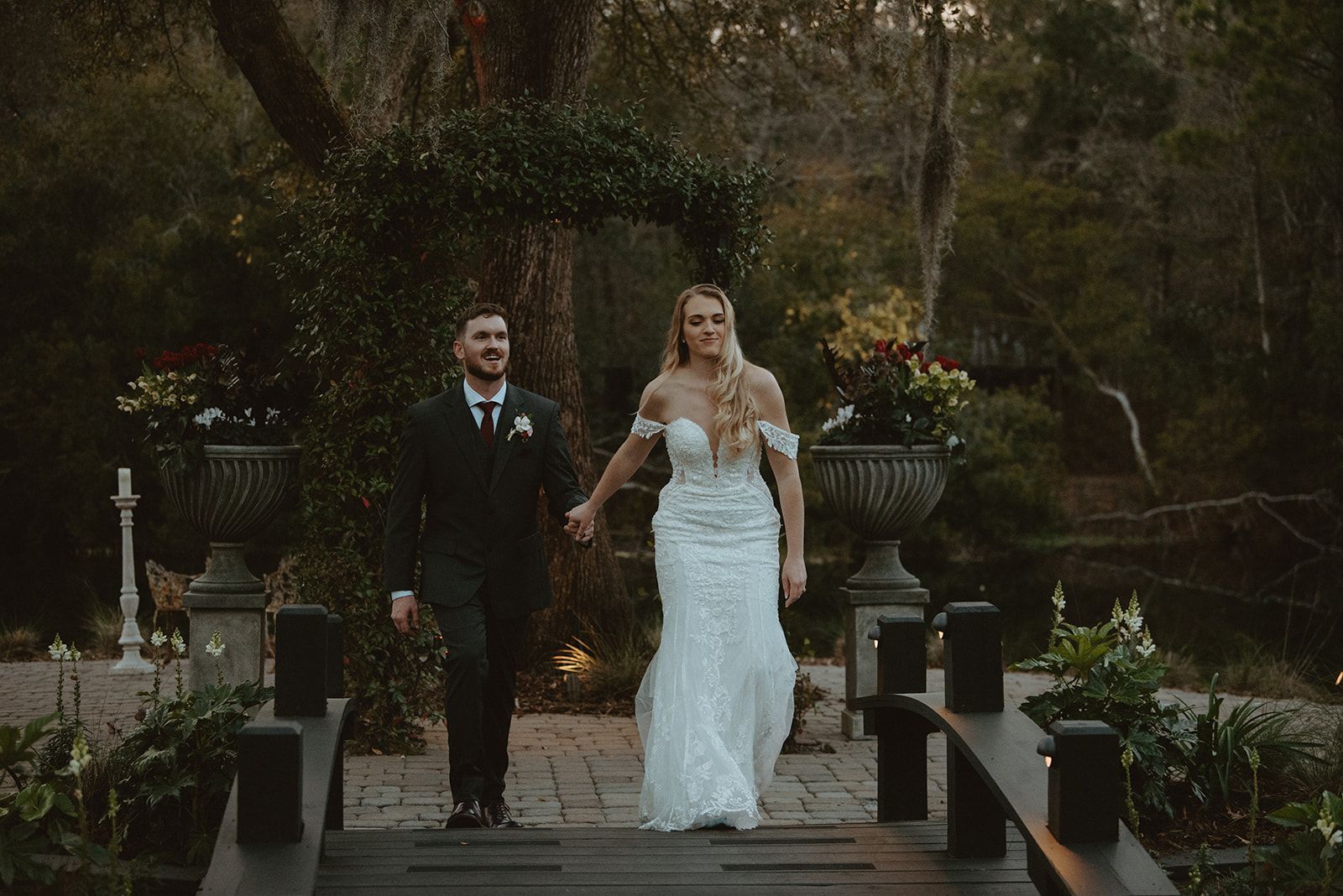 Bride and groom walking hand in hand on a garden bridge, surrounded by greenery and lanterns.