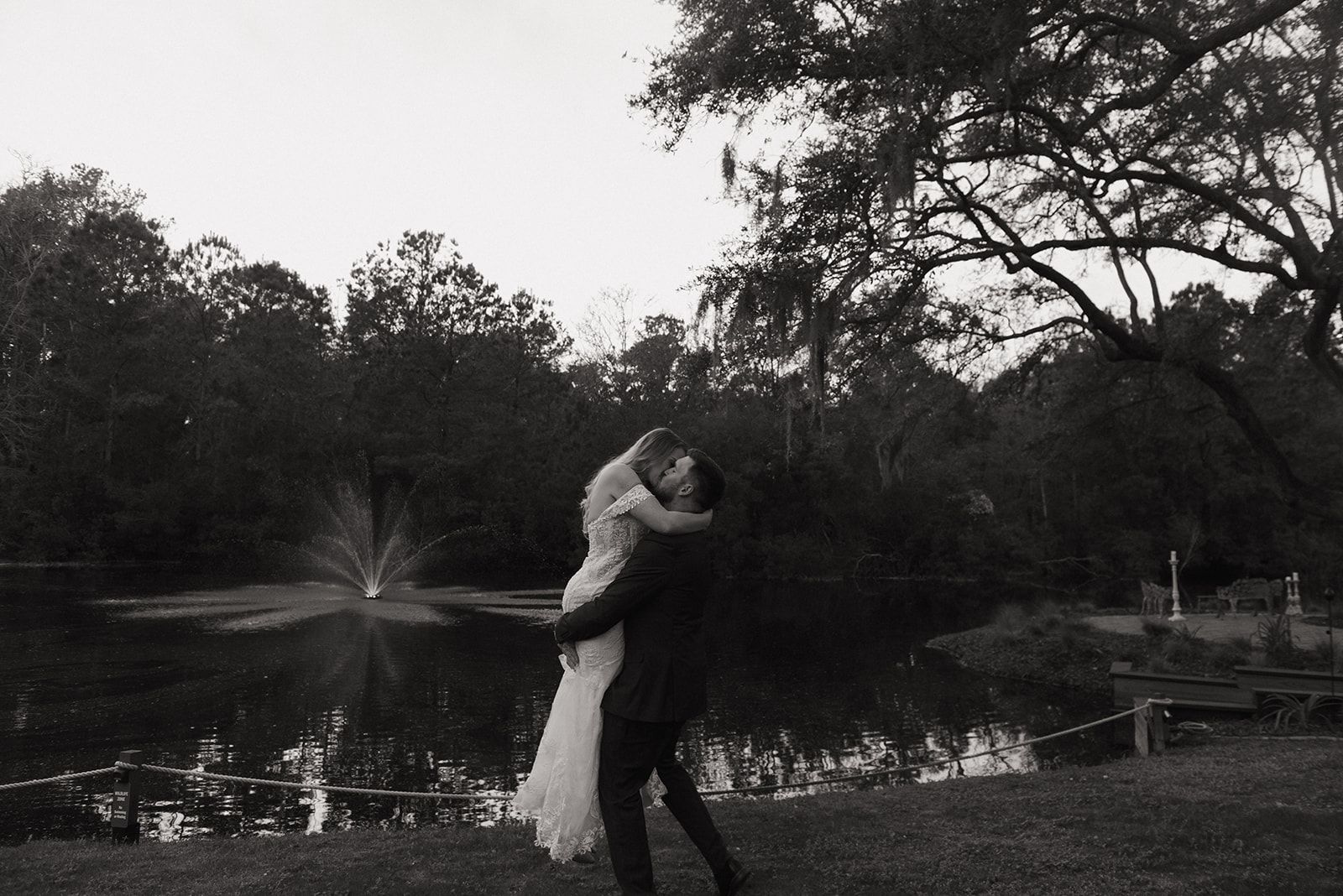 Couple embracing beside a pond with a fountain, surrounded by trees at dusk