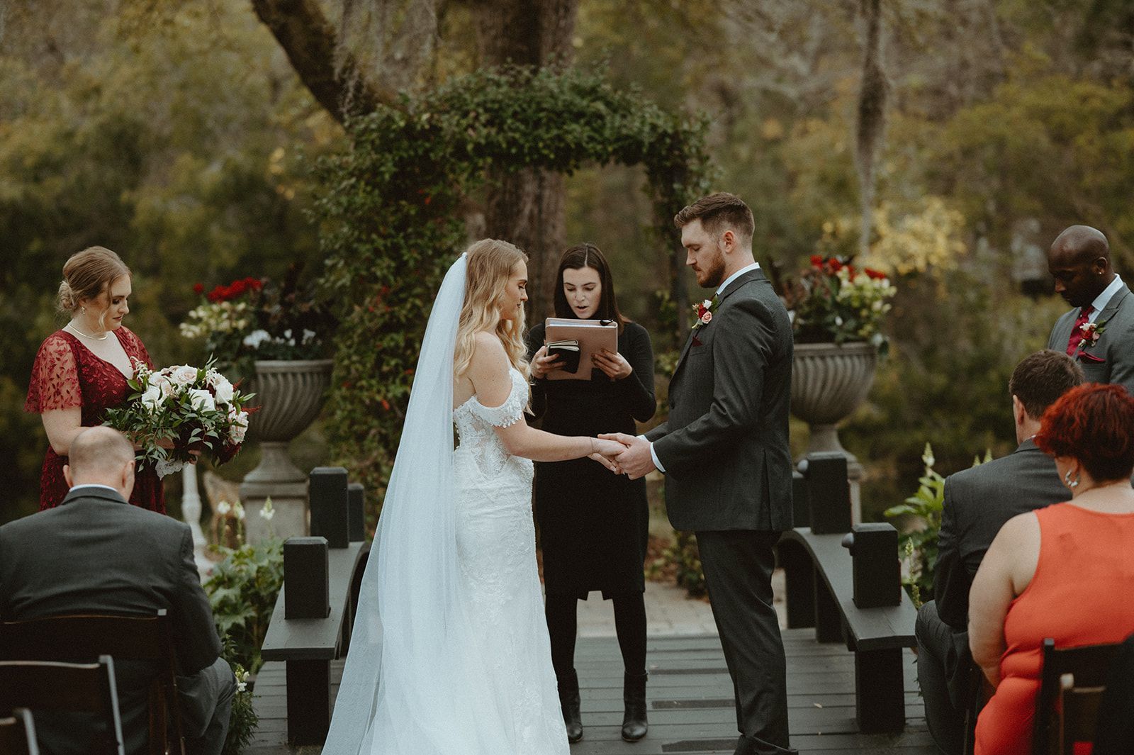 Outdoor wedding ceremony with bride and groom holding hands under an arch as an officiant stands between them