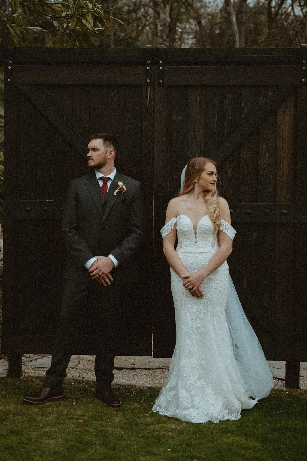 Bride in a white lace gown and groom in a dark suit stand apart before large wooden doors outdoors.